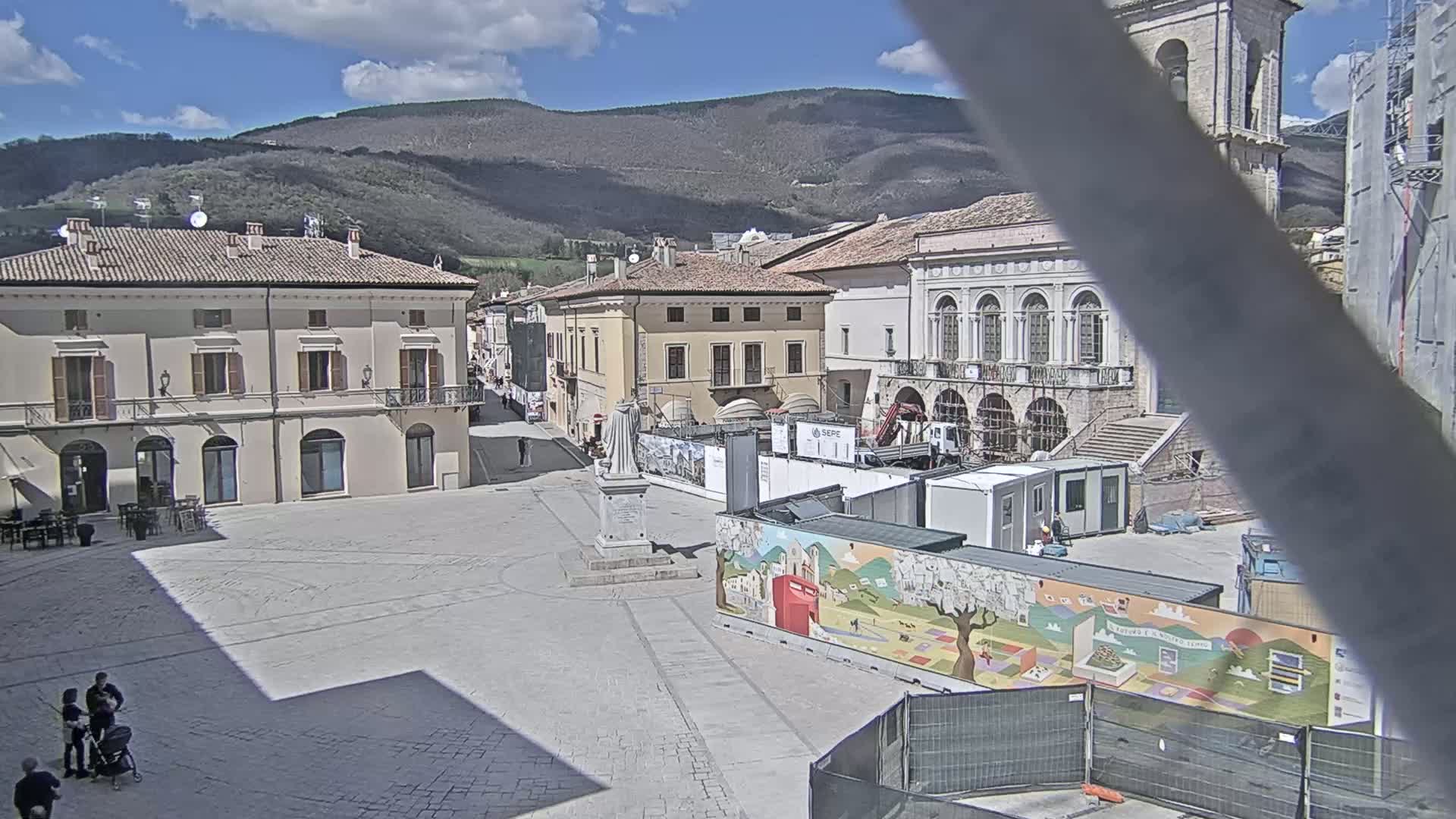 A town square with several buildings, a statue, and construction underway, all under a partly cloudy sky with mountains in the background.