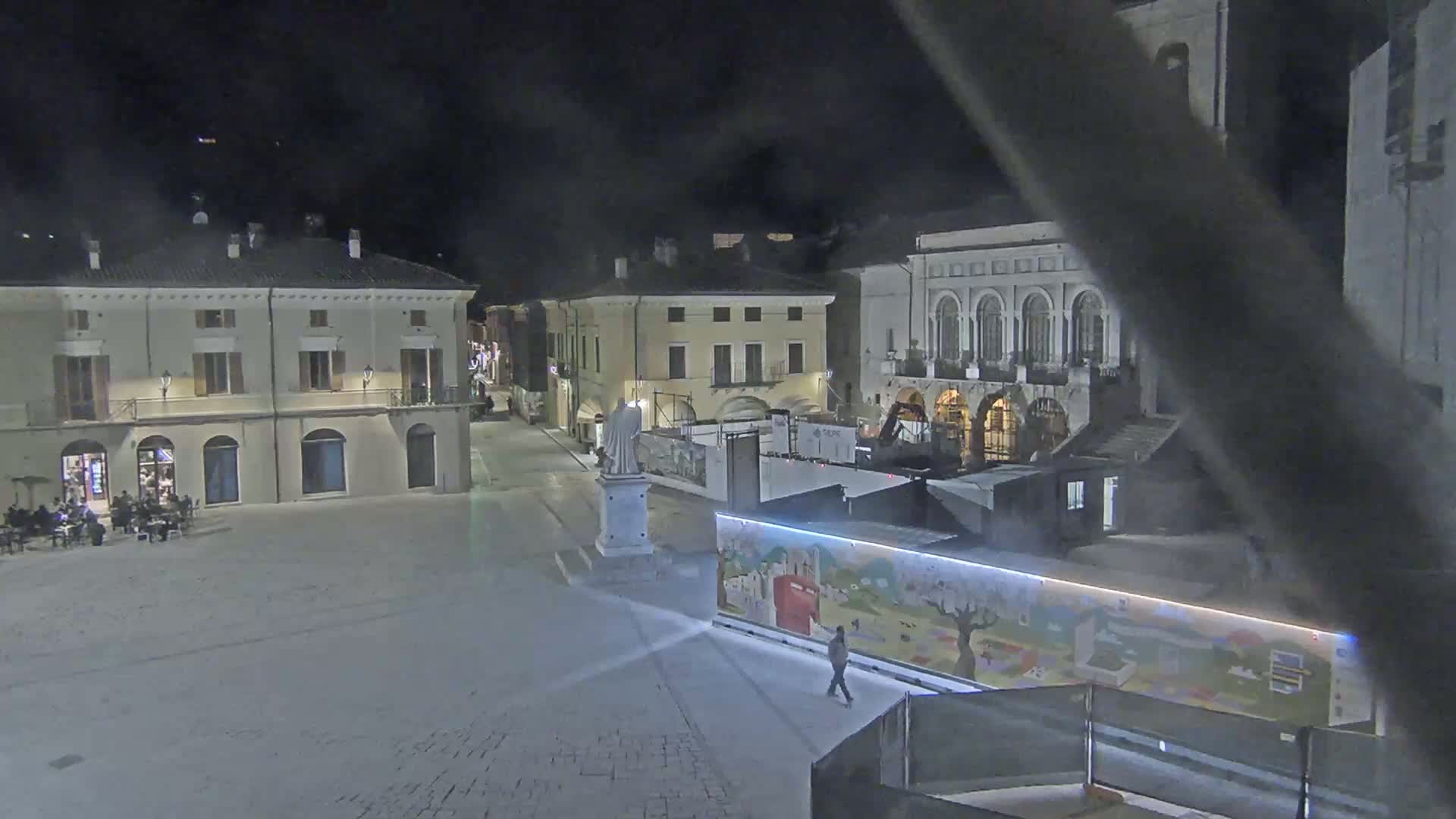 A nighttime view of a town square with several buildings, a statue, and a person walking past a brightly colored wall.