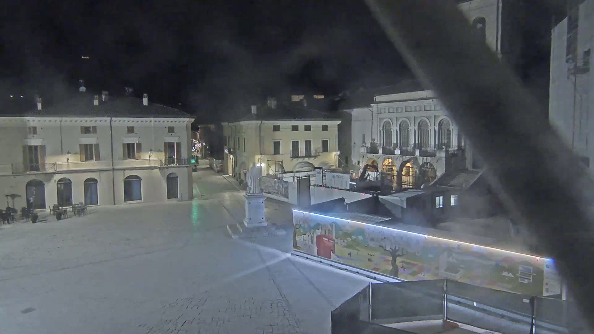 A mostly empty town square at night, with several buildings surrounding it, under hazy conditions.