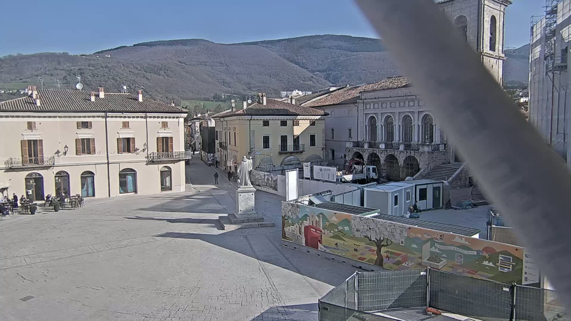 A town square with several buildings, a statue, and construction barriers under a sunny sky with mountains in the background.