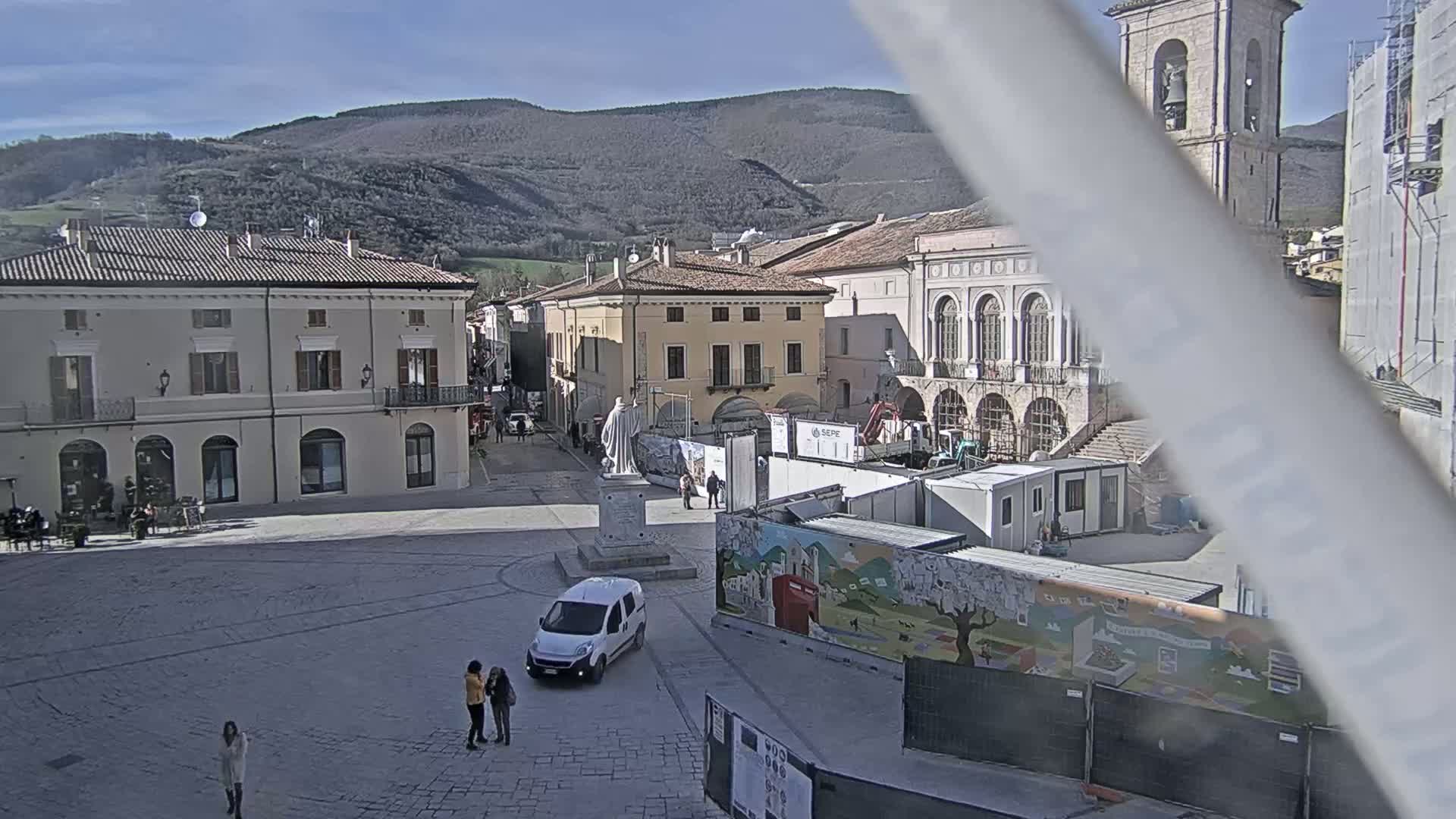 A sunny town square with several buildings, a statue, a white van, and a few pedestrians, set against a backdrop of mountains.