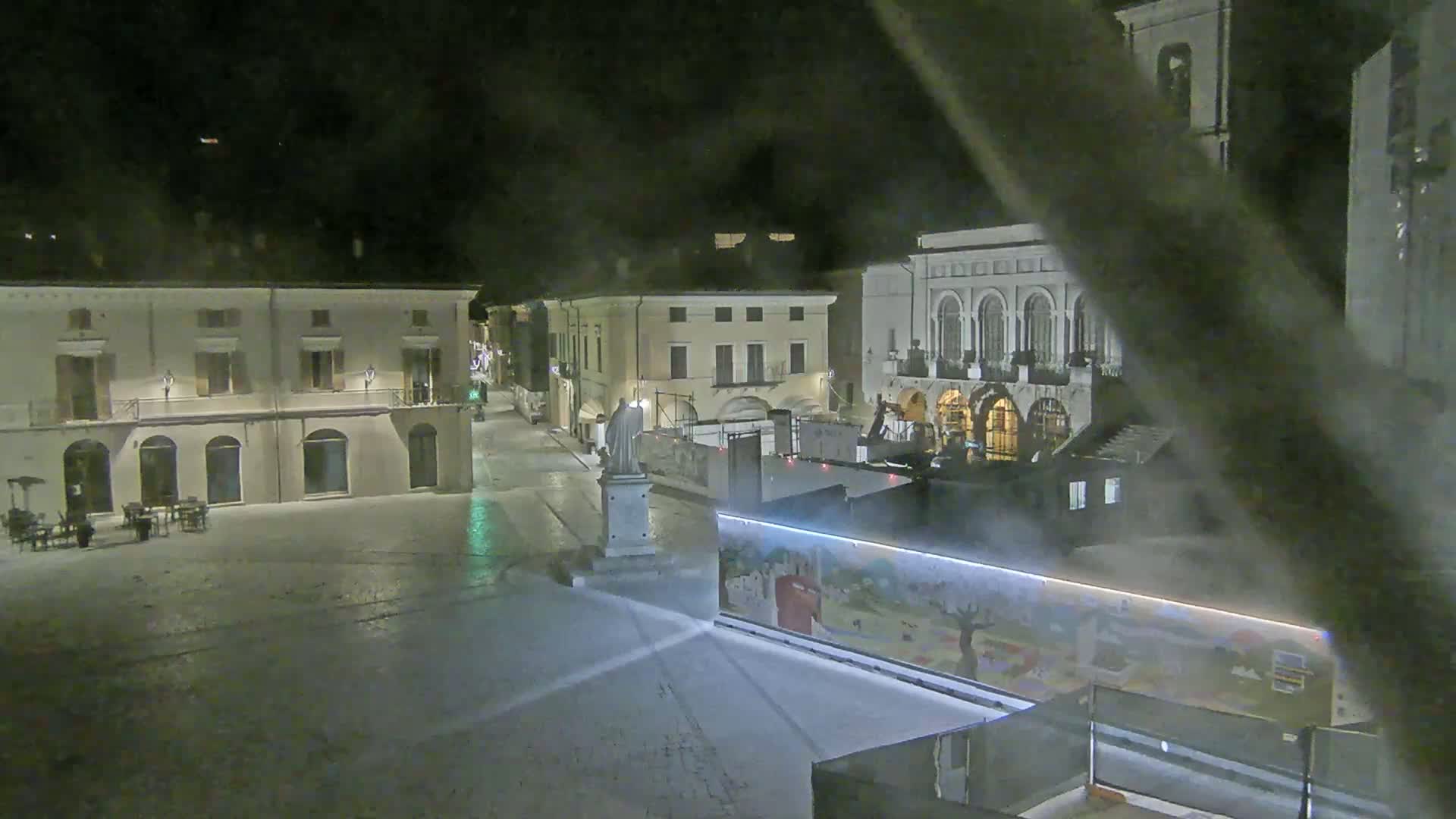 A snow-covered town square at night, featuring several buildings, a statue, and construction equipment.