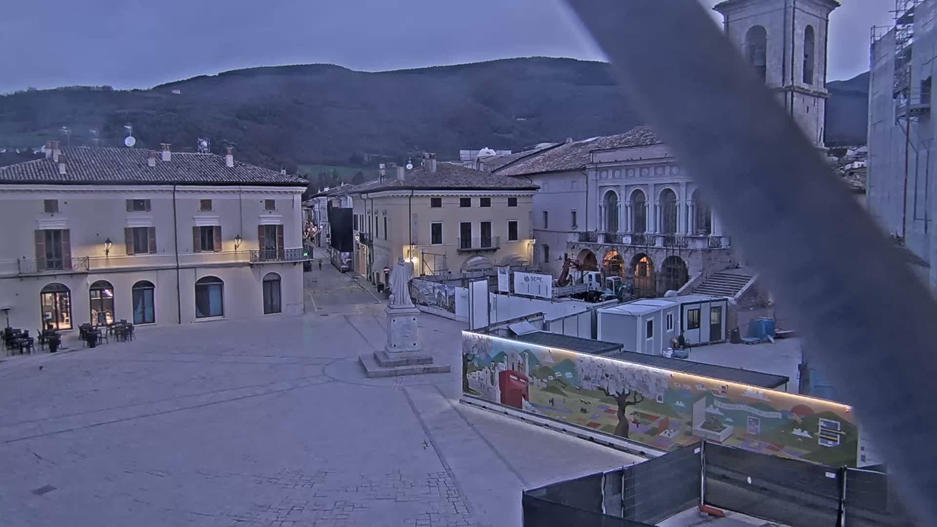 A town square with several buildings, under a dusky sky, is partially obstructed by construction equipment and barriers.