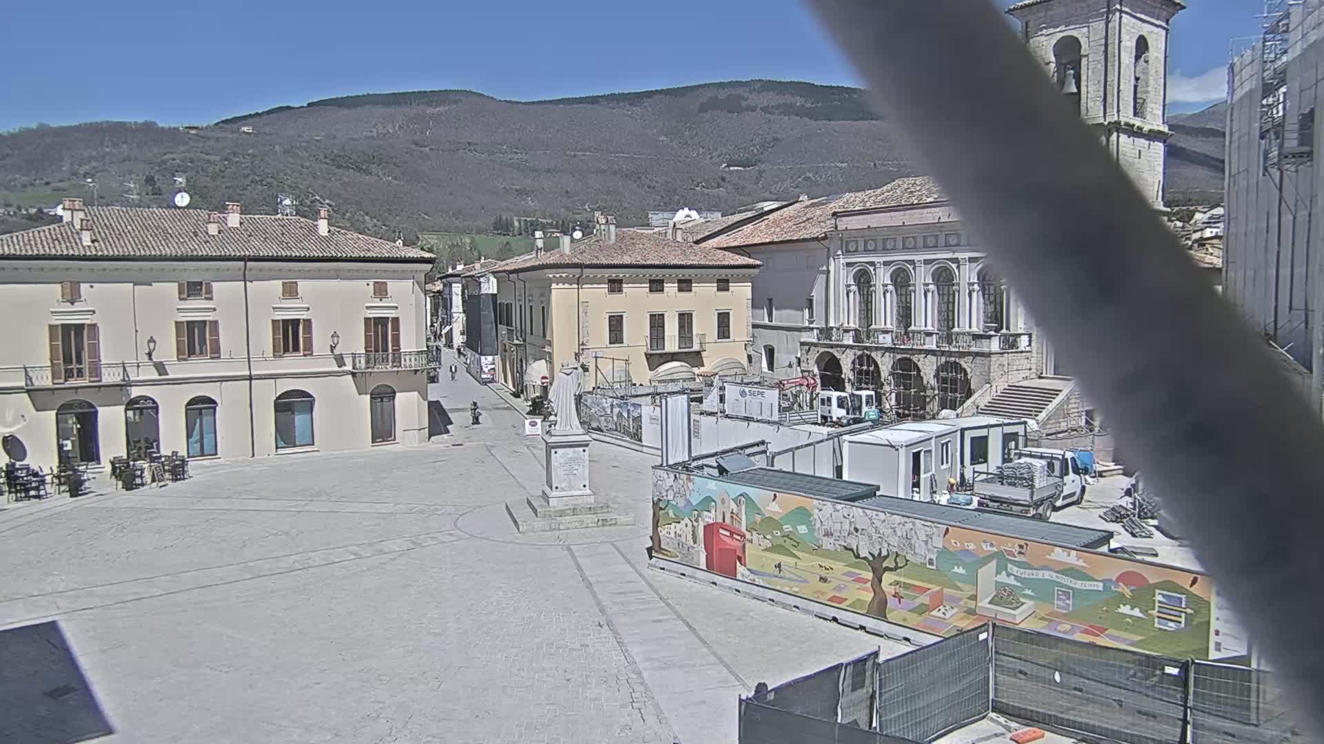 A mostly empty town square with several buildings, a statue, and construction equipment is visible under a clear, sunny sky with mountains in the background.