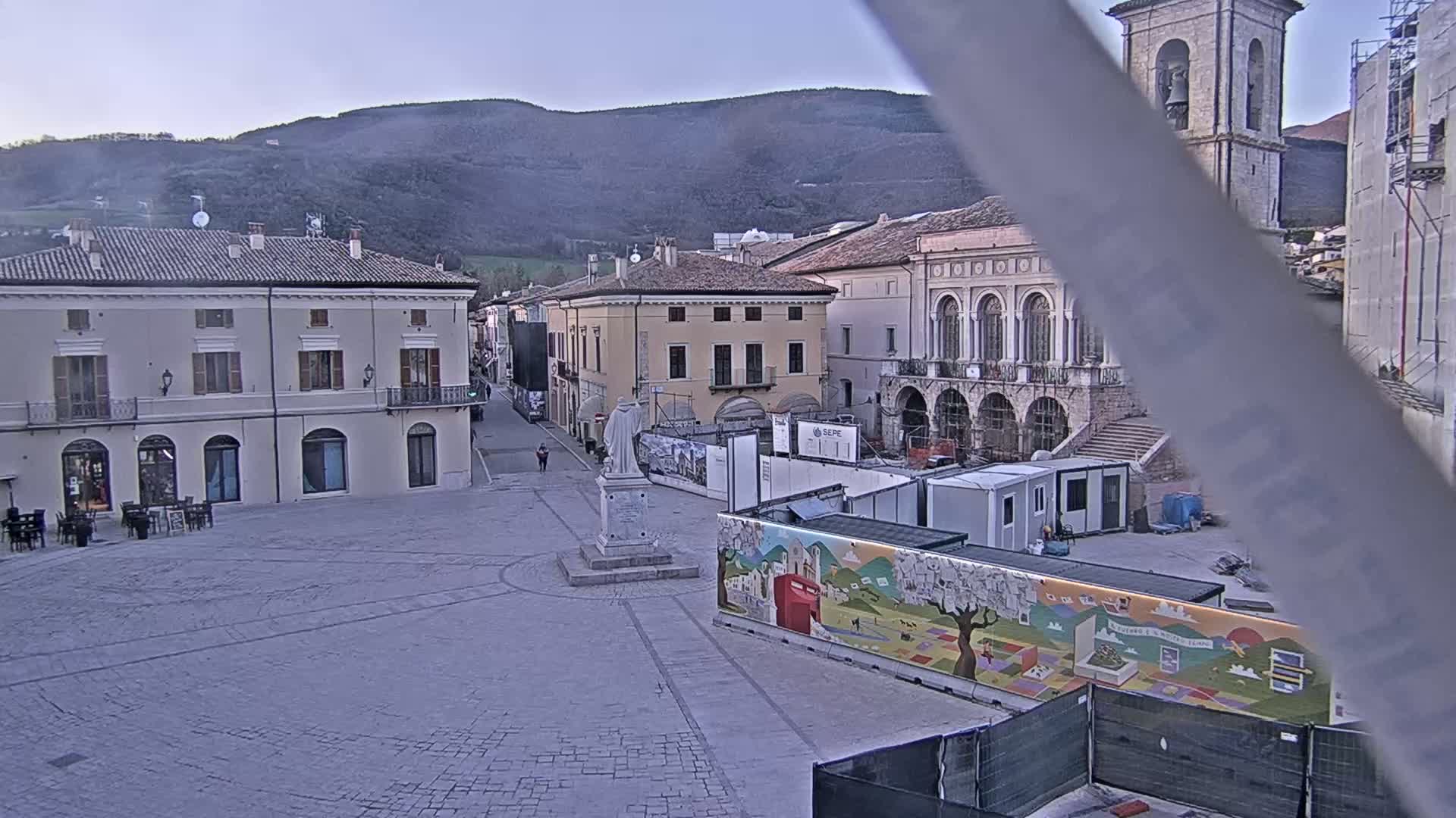 A mostly empty cobblestone town square is under construction, with buildings and a statue visible under a mostly clear, dusky sky, and mountains in the background.