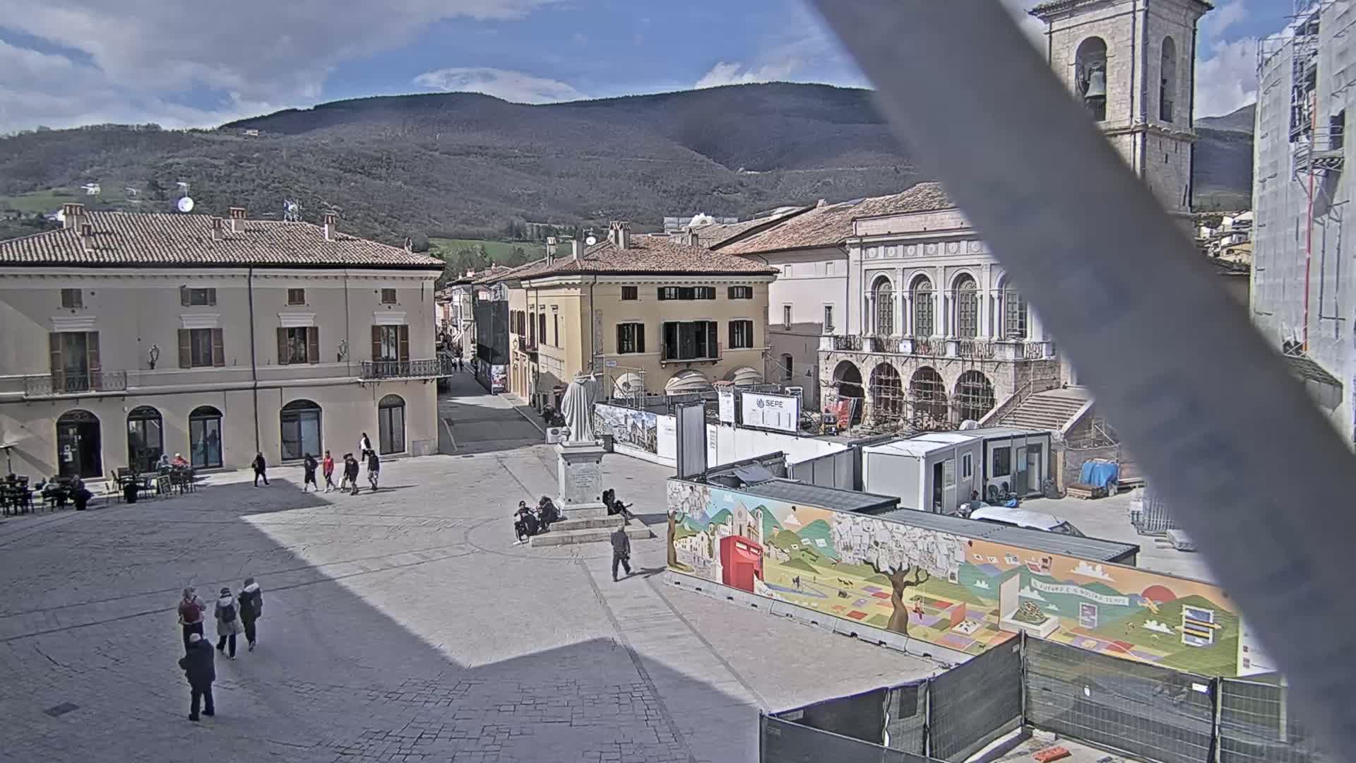 A town square under a partly cloudy sky is surrounded by buildings, some under construction, with a few people walking around.