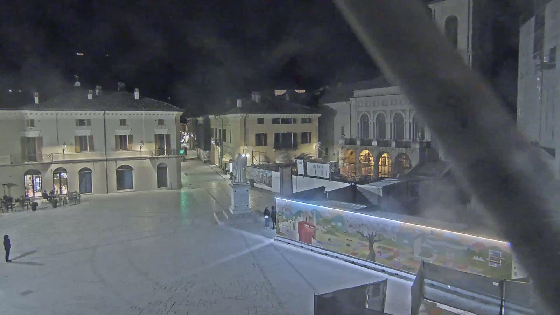 A nighttime view of a town square with several buildings, a statue, and a few people, under hazy or foggy conditions.