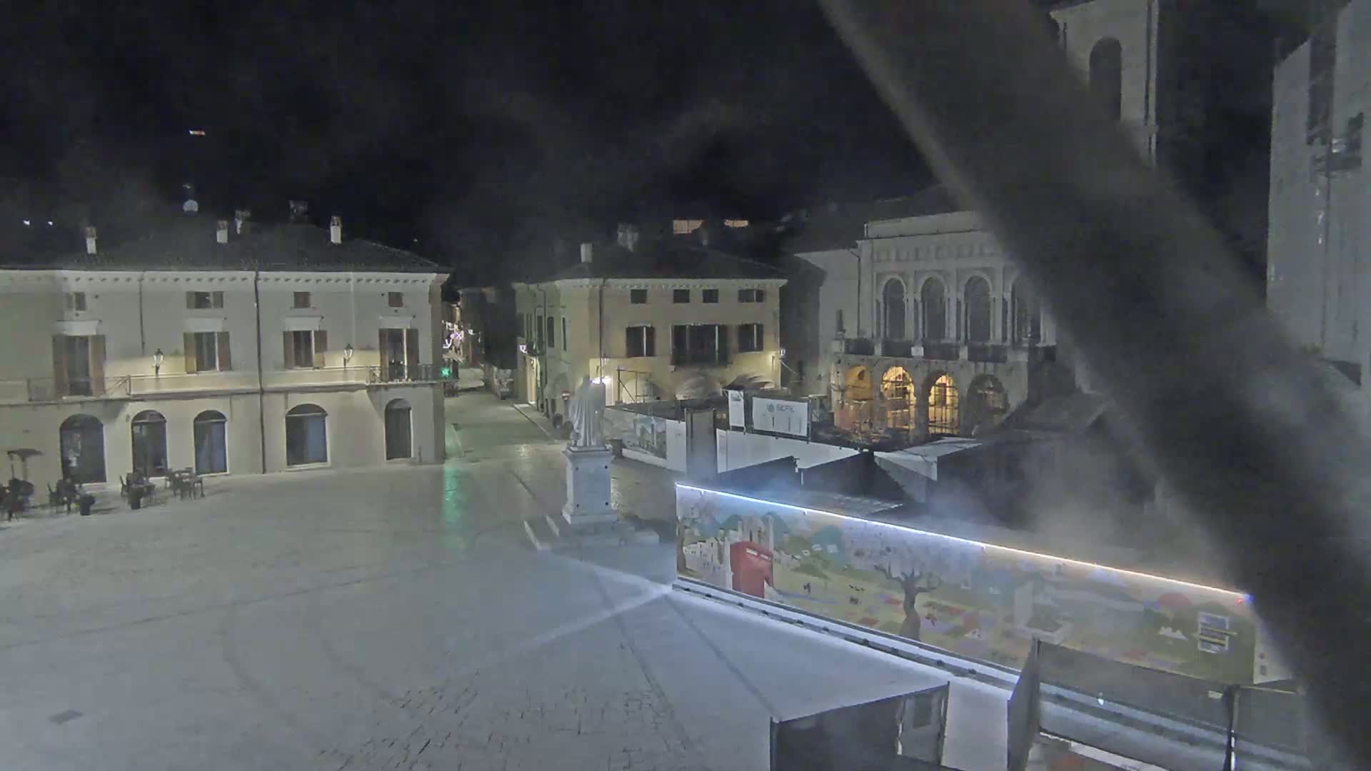 A nighttime view of a town square with several buildings surrounding it, partially obscured by fog or mist.