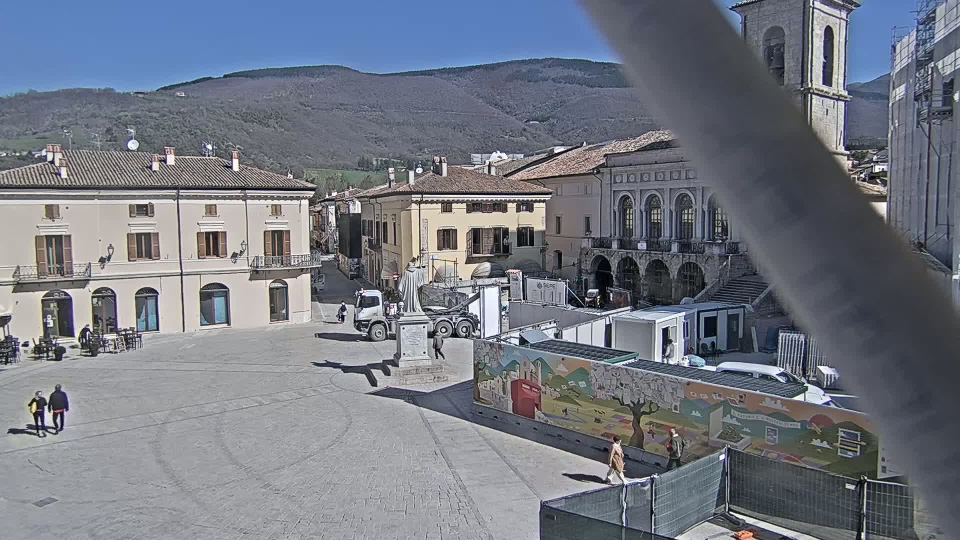 A town square, under sunny skies, features several buildings, a statue on a pedestal, construction equipment, and a few pedestrians.