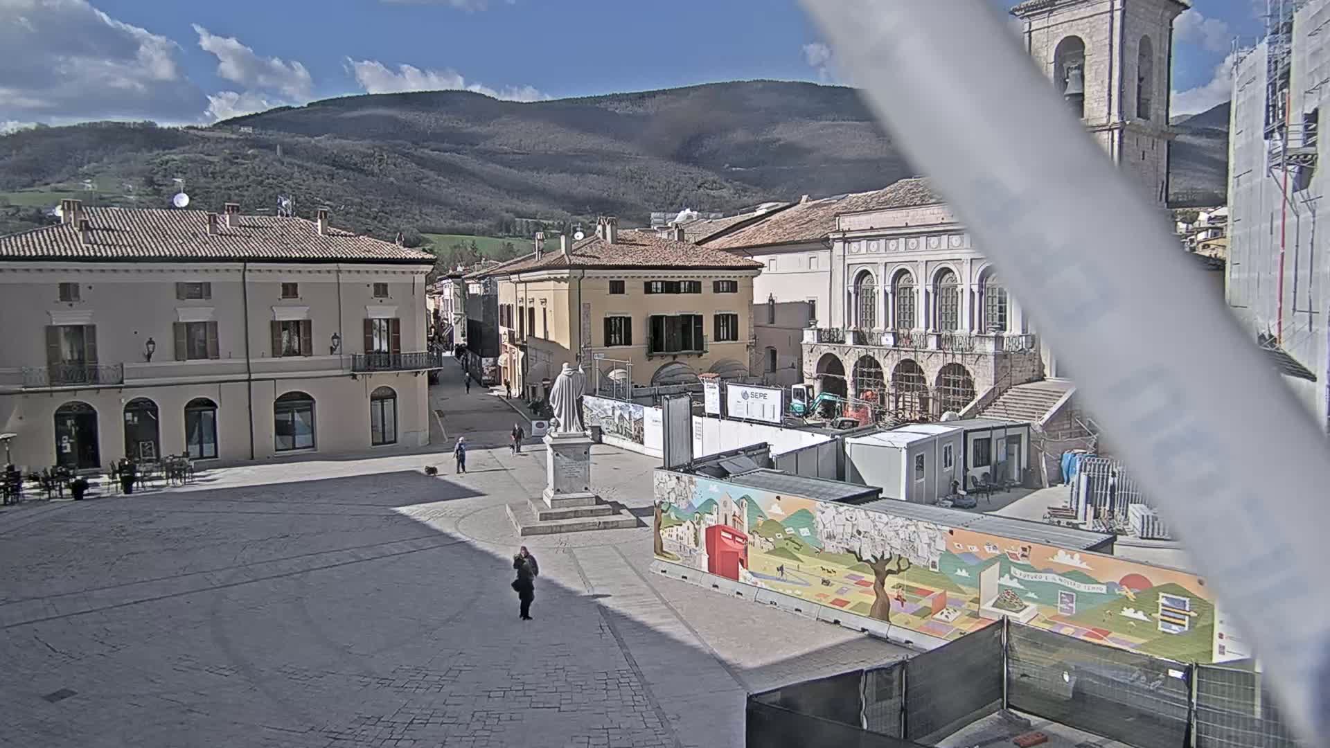 A town square in a sunny, mountainous region, shows construction underway near a statue and historical buildings.