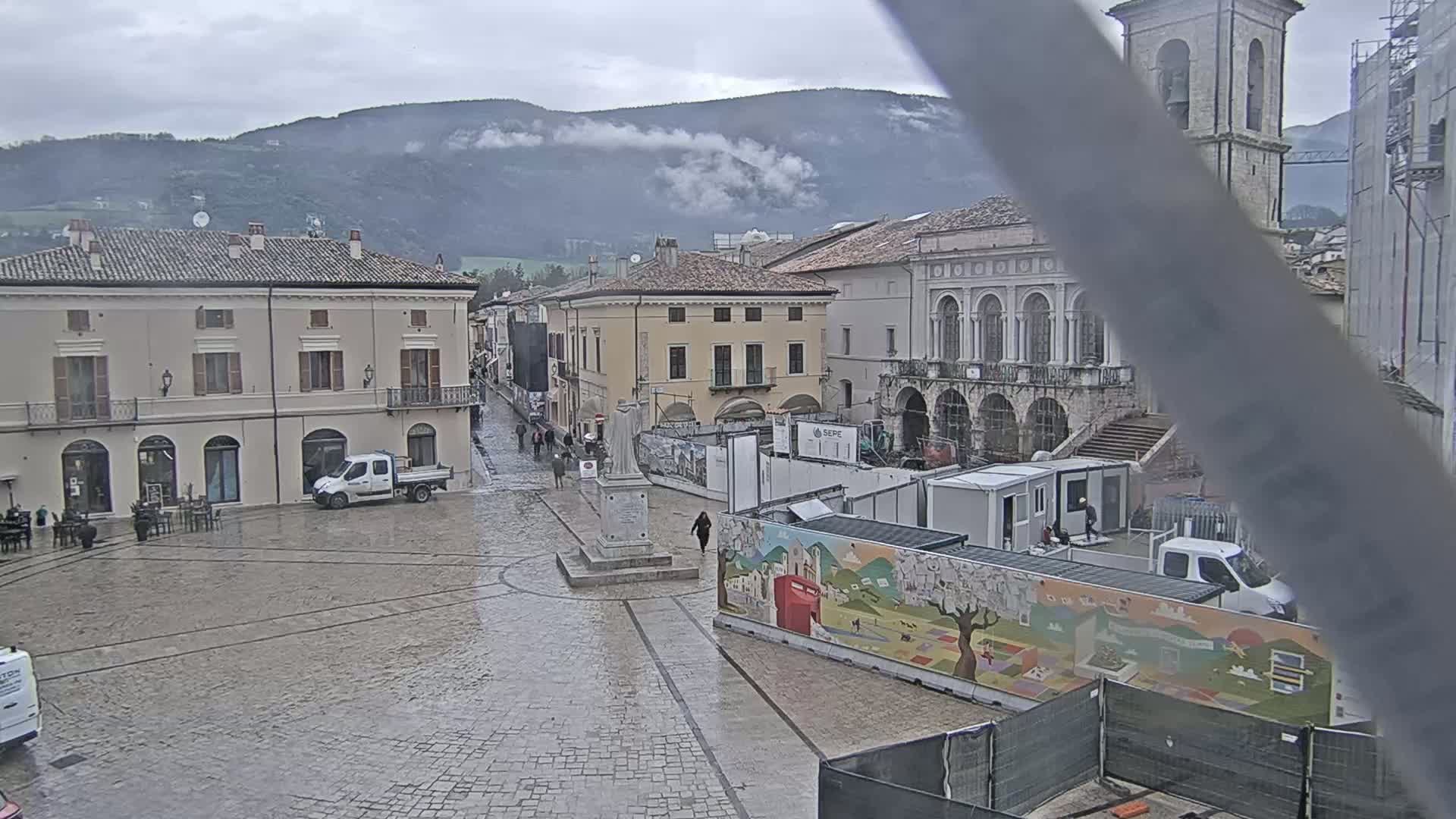 A wet, stone plaza in a town nestled in the foothills of misty mountains features several buildings, construction equipment, and a few pedestrians.