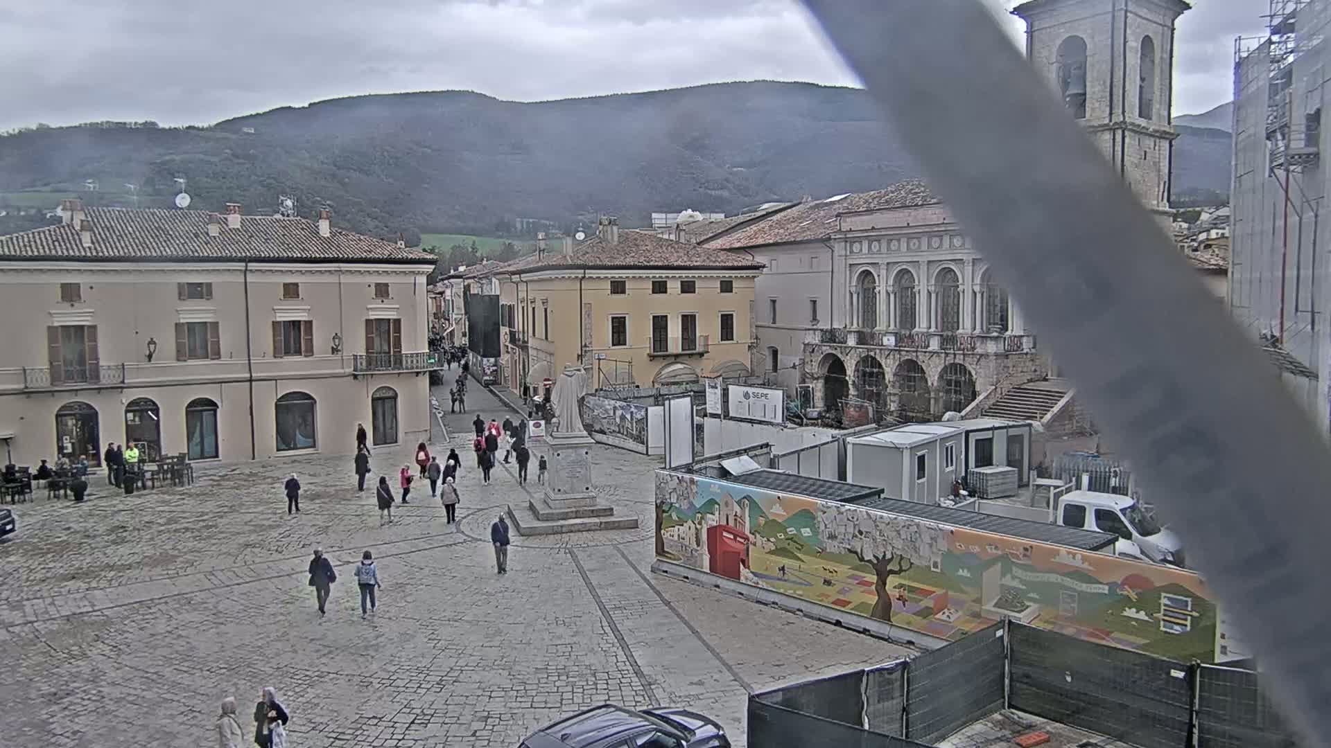A cobblestone town square with several people walking around, surrounded by buildings, under an overcast sky with mountains in the background.