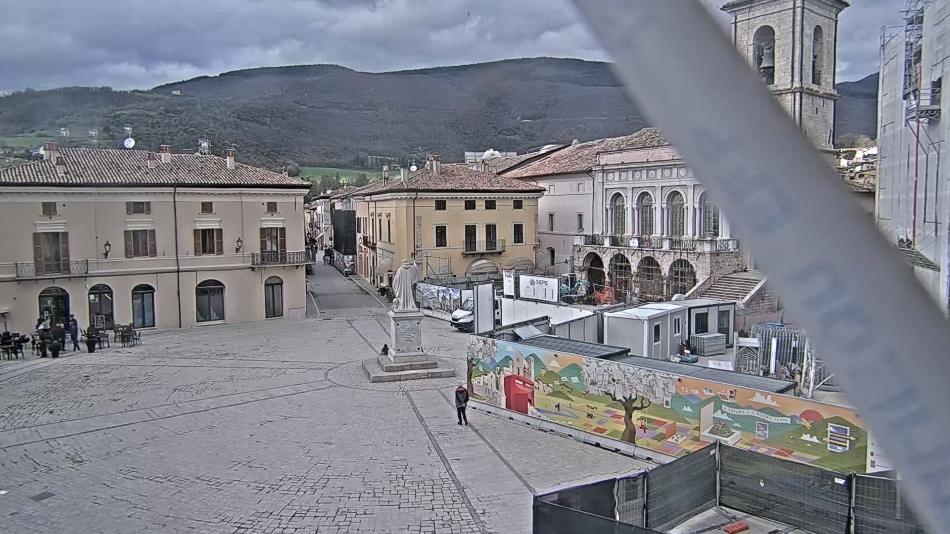 A town square in a city nestled in a valley under a cloudy sky features several buildings, construction work, and a statue in the center.