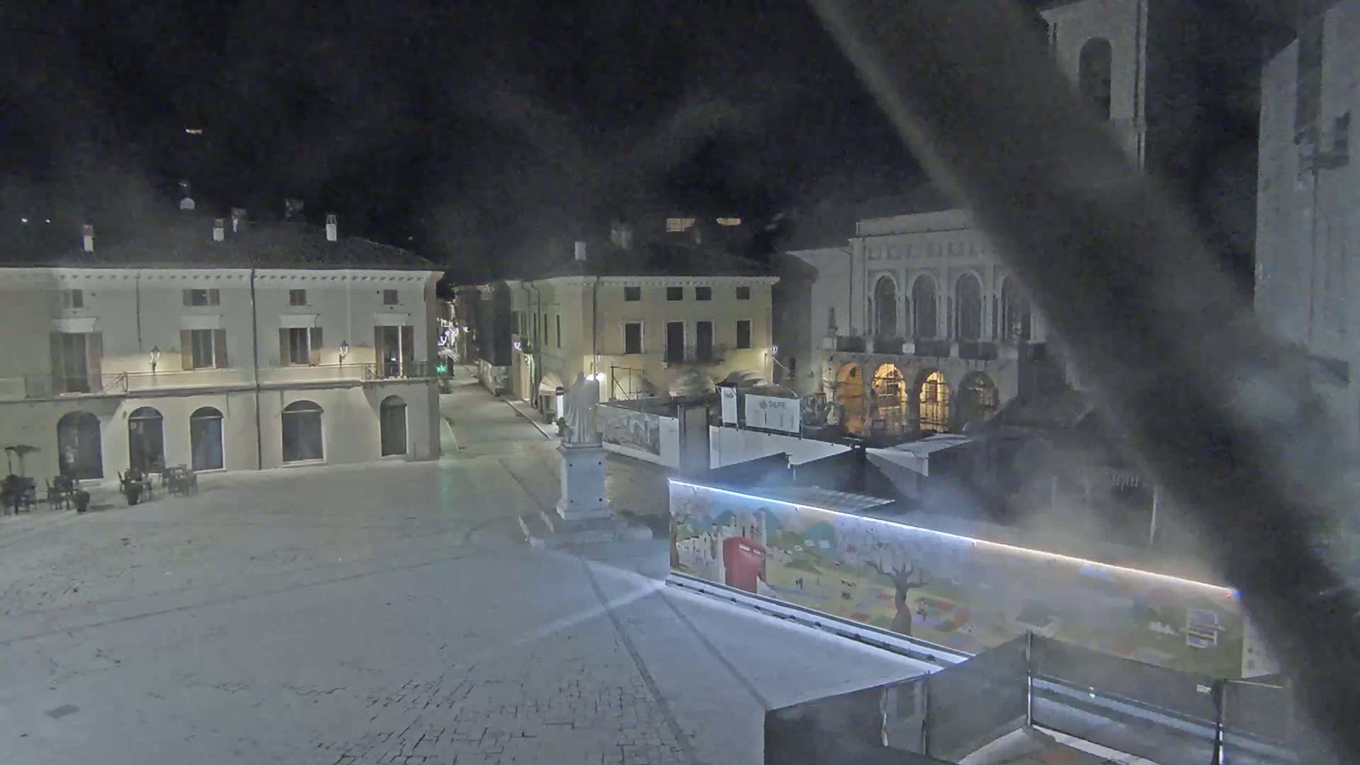 A snow-covered town square at night, illuminated by streetlights, with several buildings surrounding it and a statue in the center.