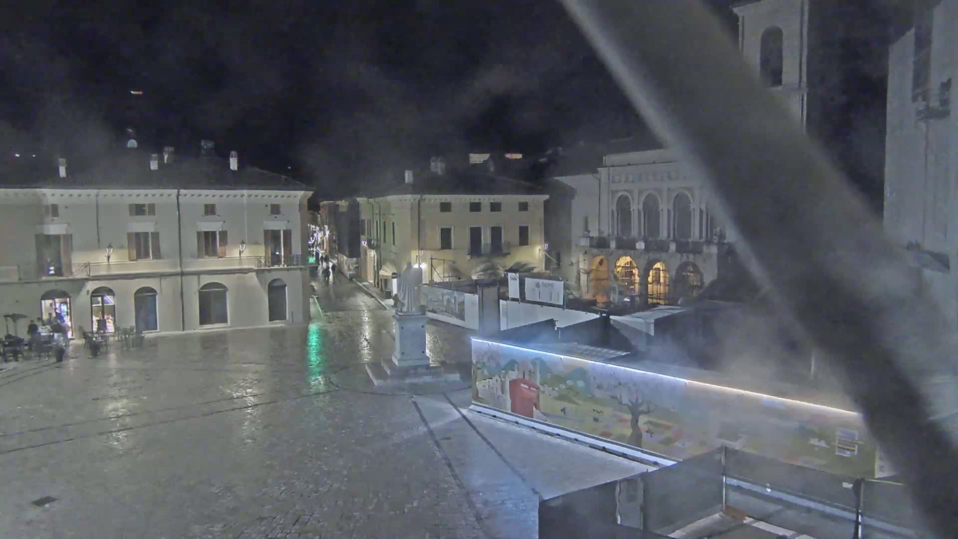 A wet, nighttime view of a town square with several buildings, a statue, and a colorful construction barrier.
