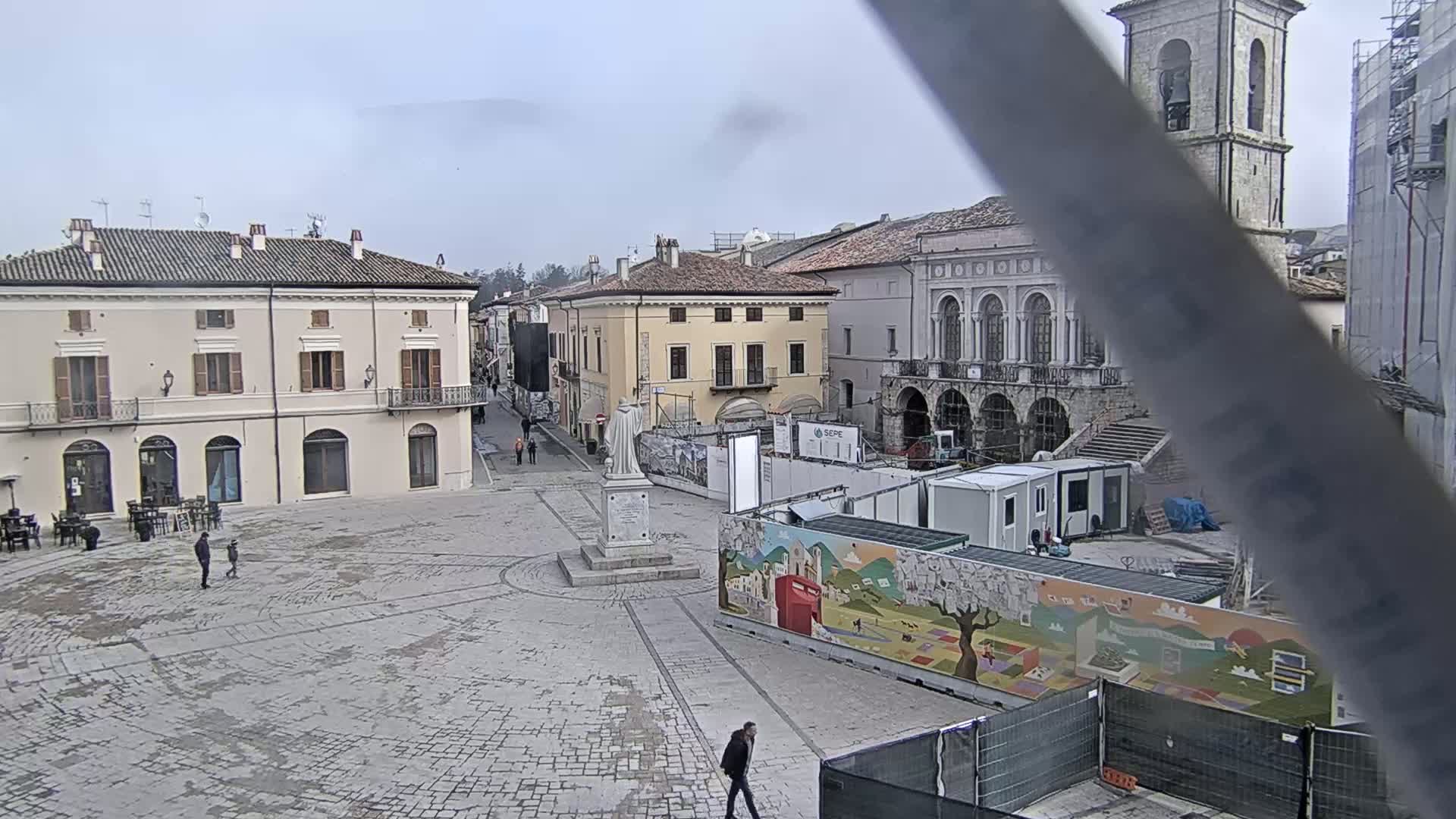 A mostly empty town square with several buildings surrounding it, under an overcast sky, shows ongoing construction near the center of the square.