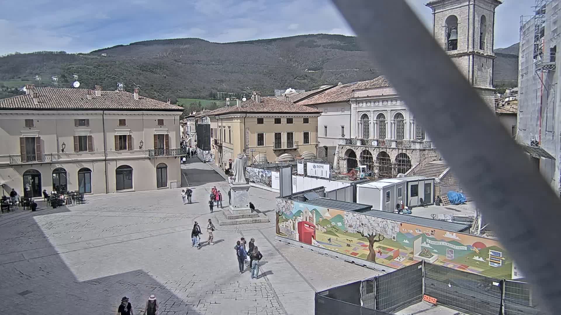 A town square with several buildings, a few pedestrians, and construction underway under a mostly sunny sky with mountains in the background.