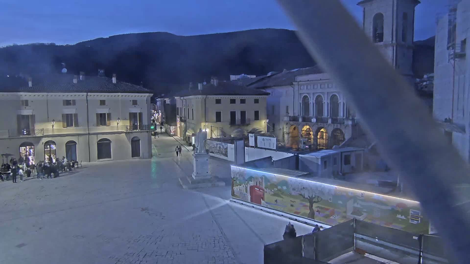 A town square at twilight, under a dark sky, is surrounded by buildings and features a statue and construction barriers with a painted mural.