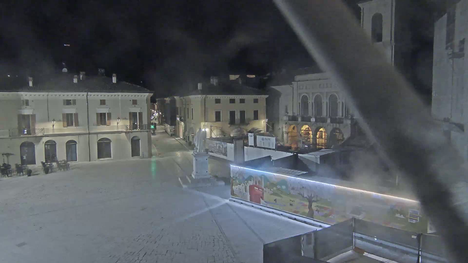 A nighttime view of a town square with several buildings, a statue, and construction barriers with artwork.