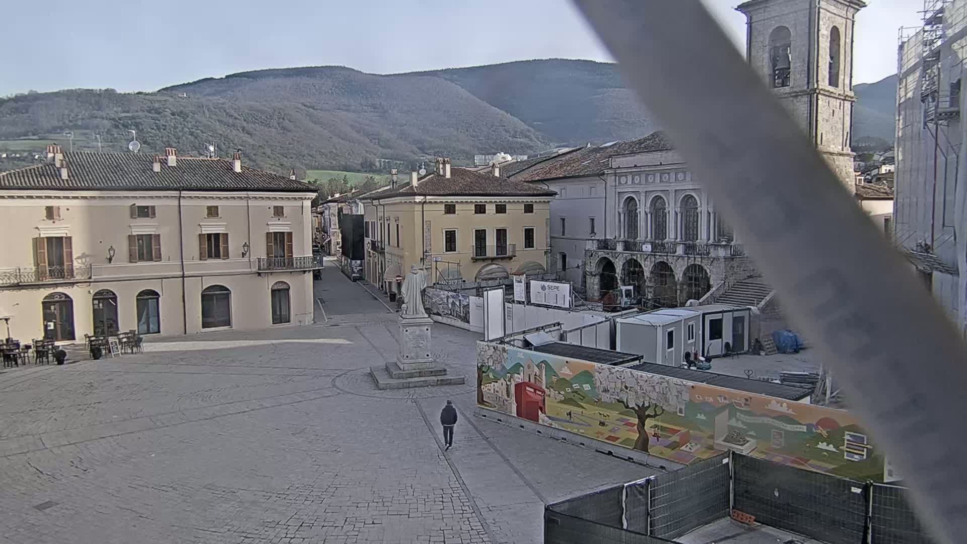 A mostly empty town square with several buildings, a statue, and construction underway, all under a clear, sunny sky with mountains in the background.