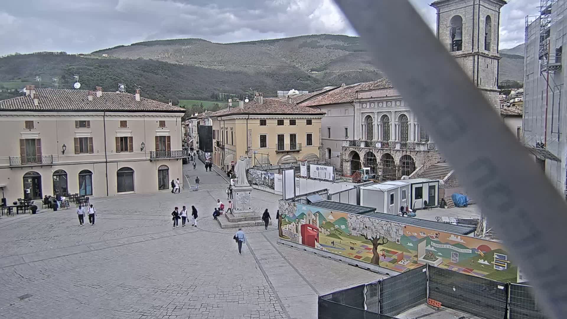 A town square with several buildings, a statue, and some people walking around under an overcast sky, with mountains visible in the background.