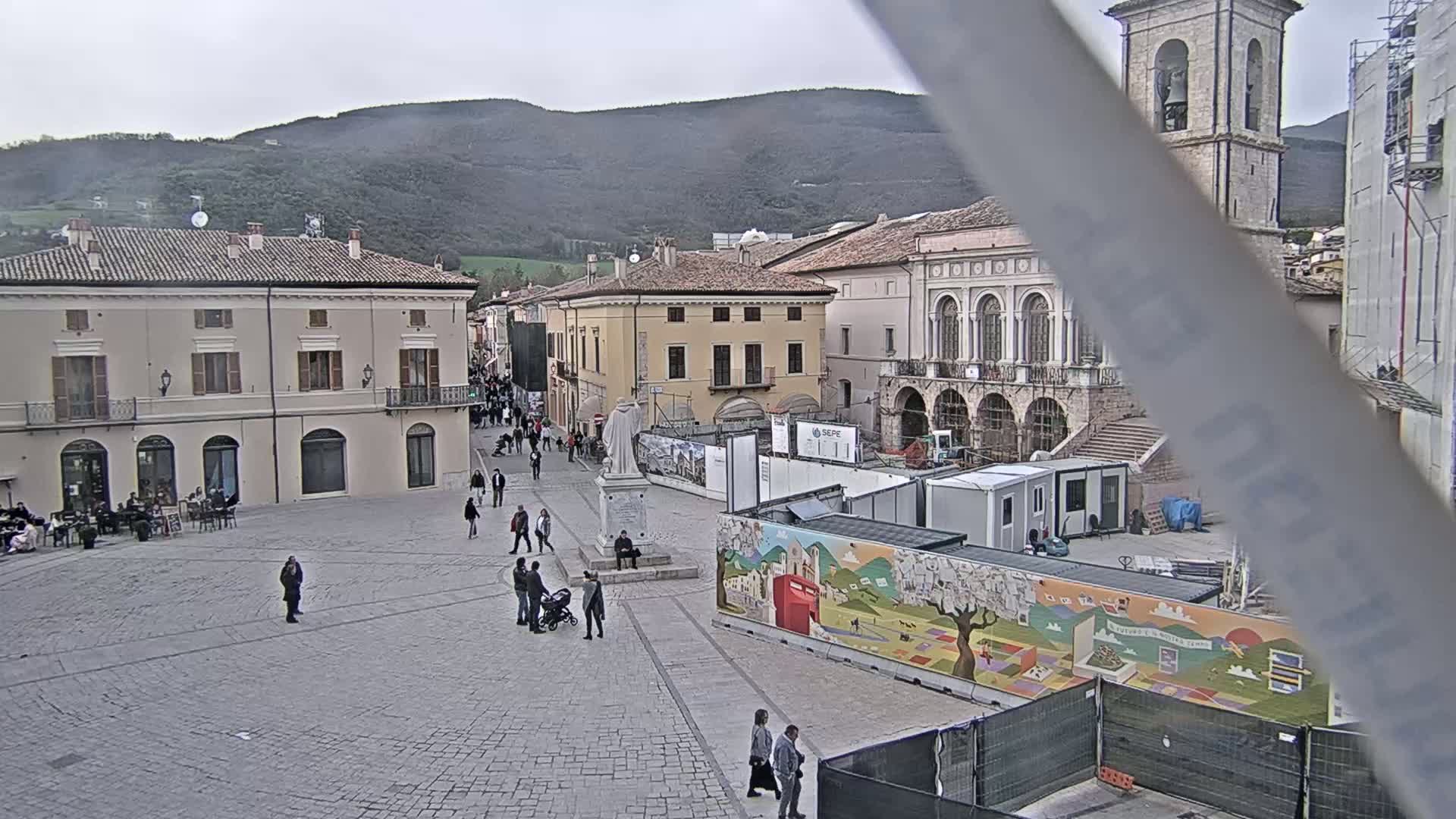 A town square with several buildings, a statue, and a few people, under an overcast sky, with construction underway in part of the square.