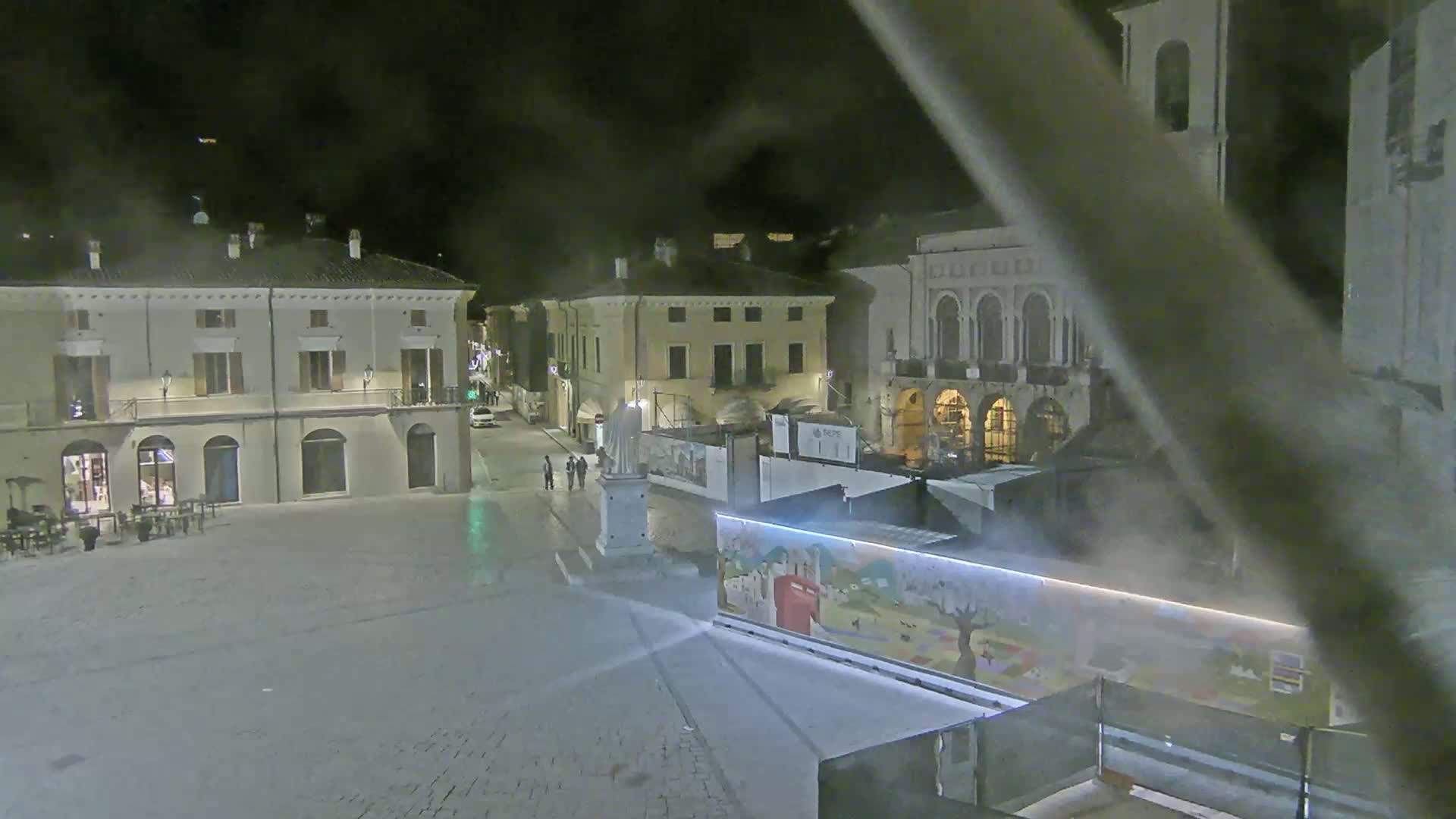 A nighttime view of a town square, featuring several buildings, a statue, and a brightly-lit structure with artwork on its side.