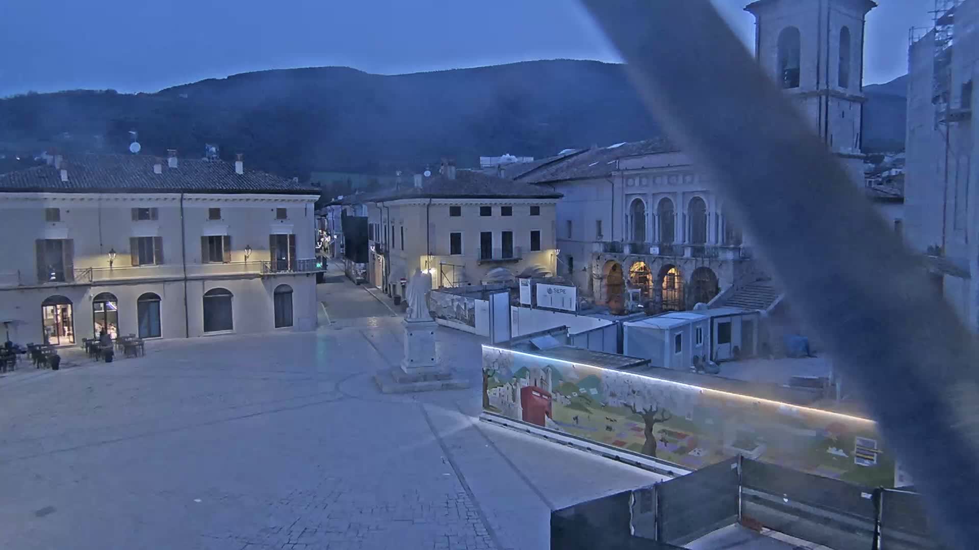 A mostly empty town square at dusk or dawn, with several buildings, a statue, and construction barriers, is set against a backdrop of mountains.