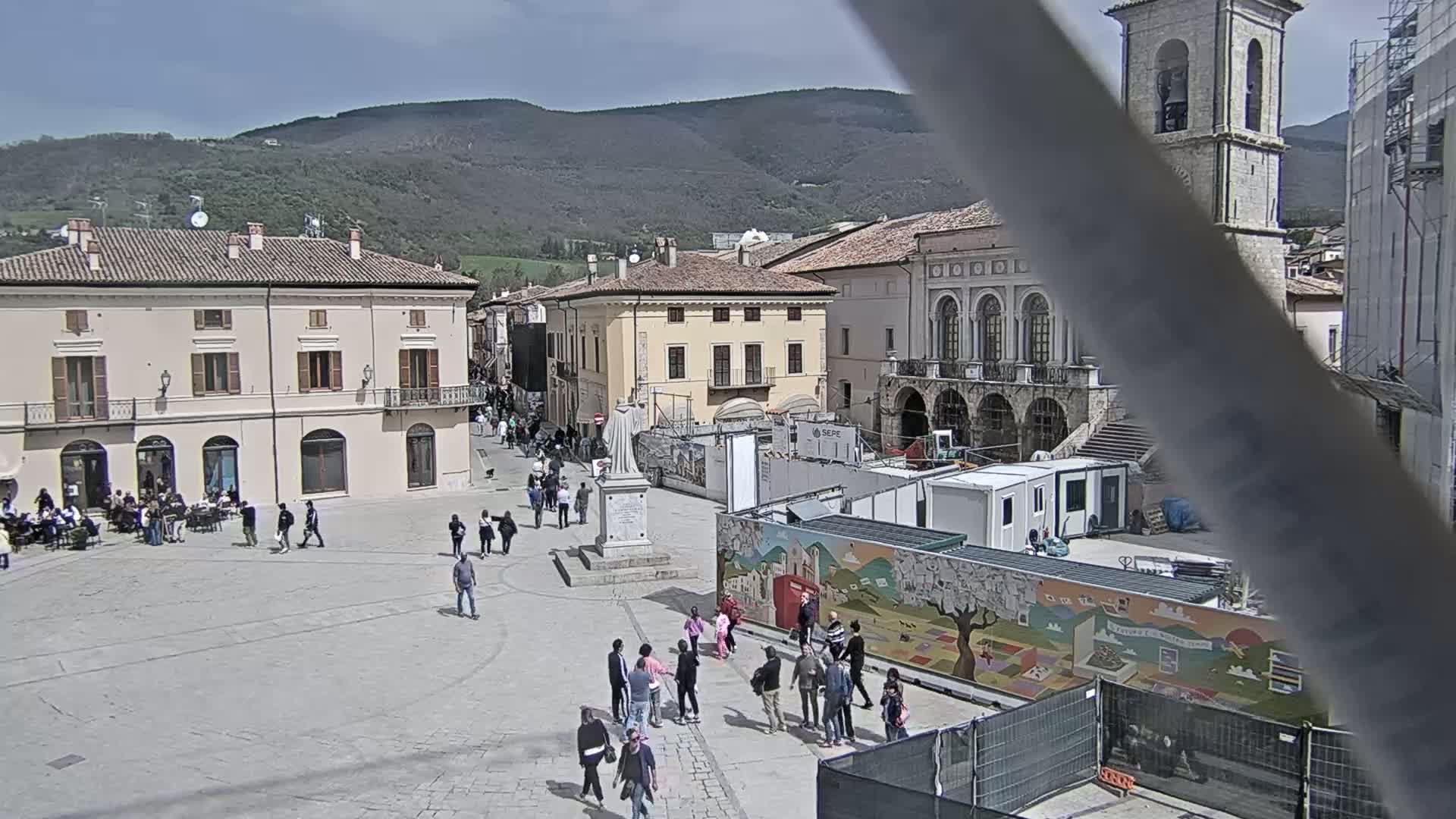 A town square, under a partly sunny sky, is populated with people, buildings, and construction.