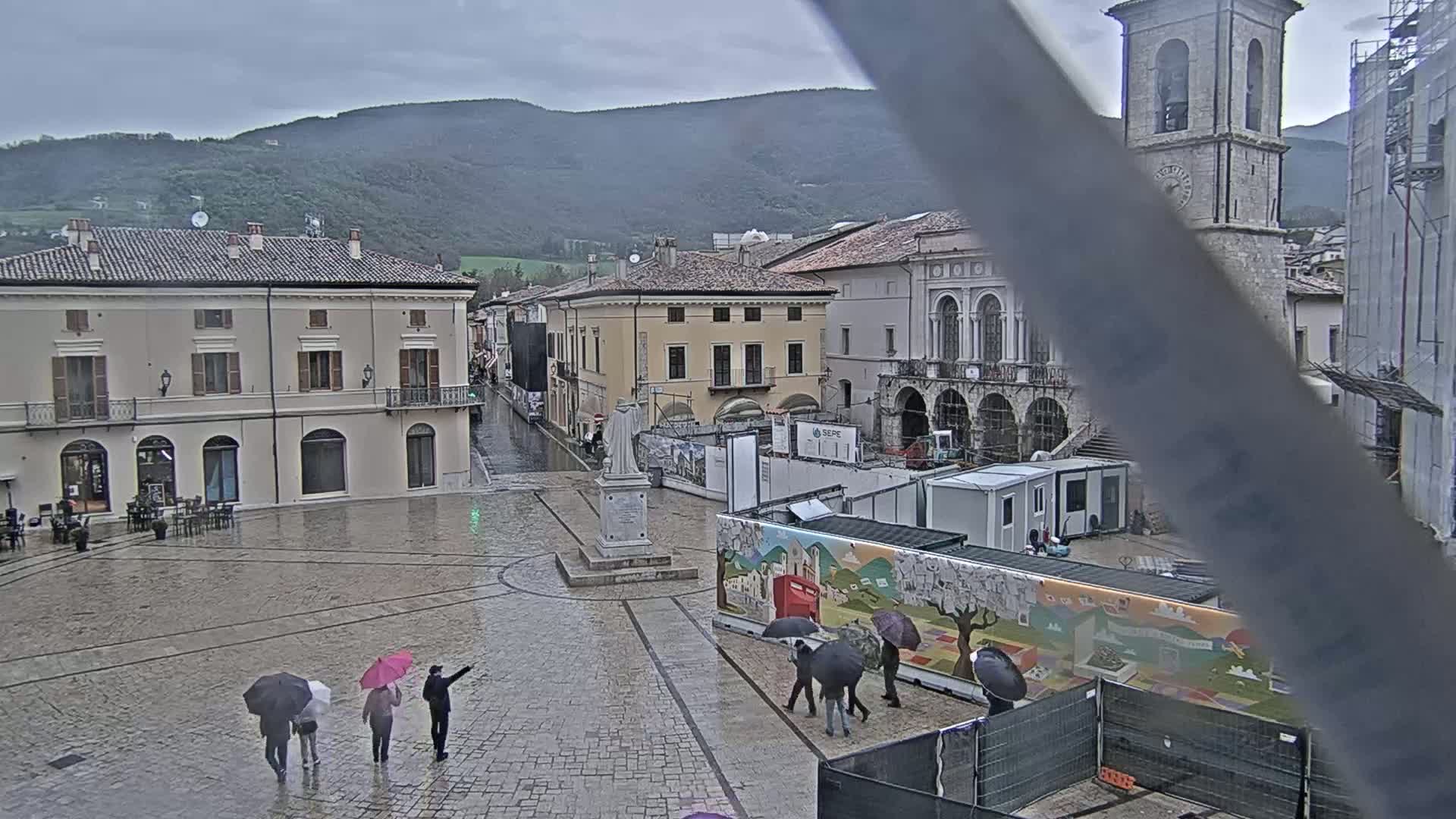 A rain-soaked town square, surrounded by historic buildings and mountains in the distance, features several people with umbrellas.