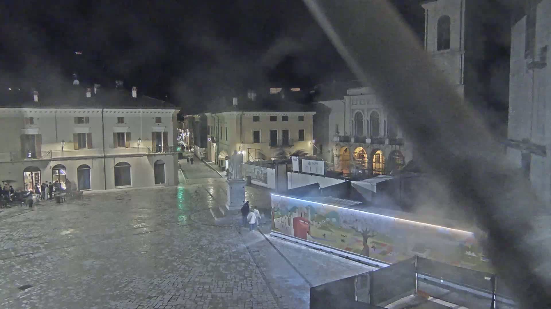 A nighttime view of a cobblestone town square, lightly misted, with several buildings surrounding it, some with lights on.