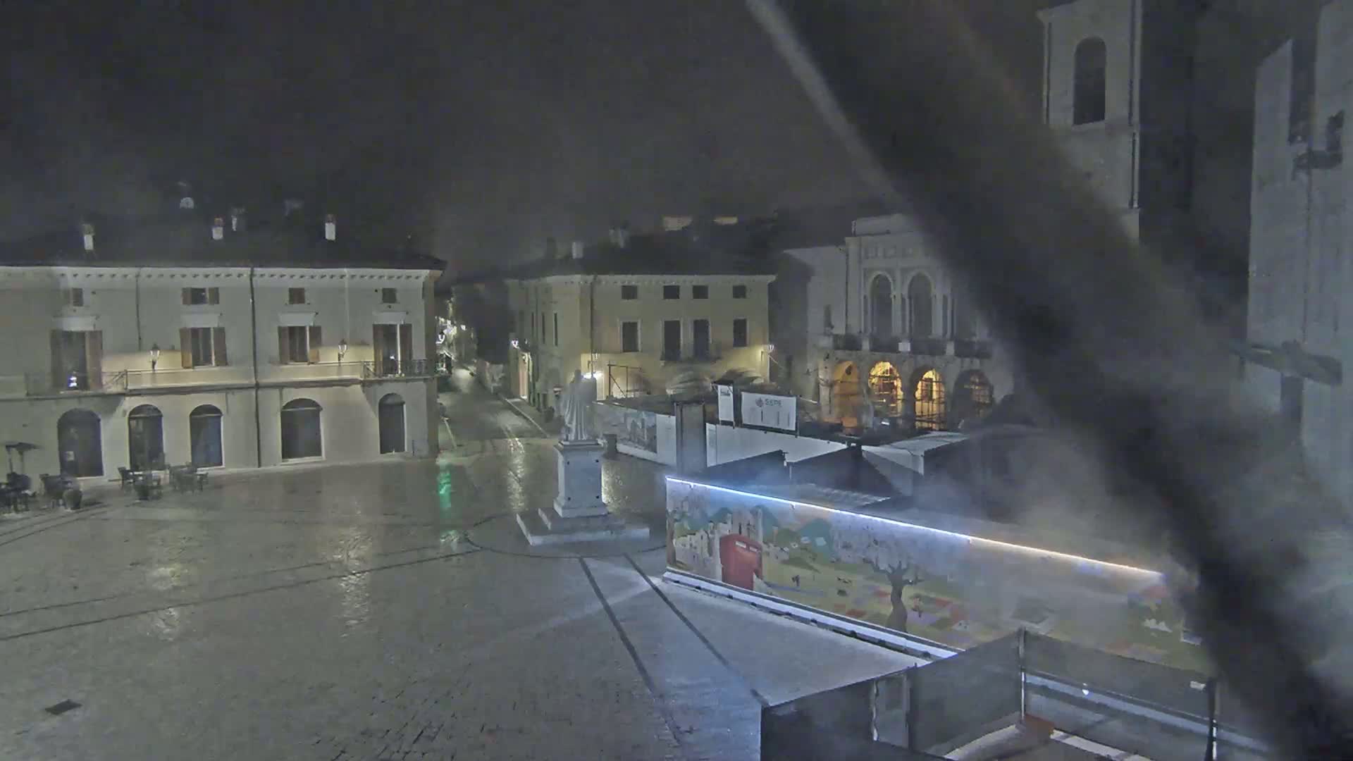 A statue stands in a mostly empty town square at night, under a hazy, possibly rainy sky, surrounded by several buildings.