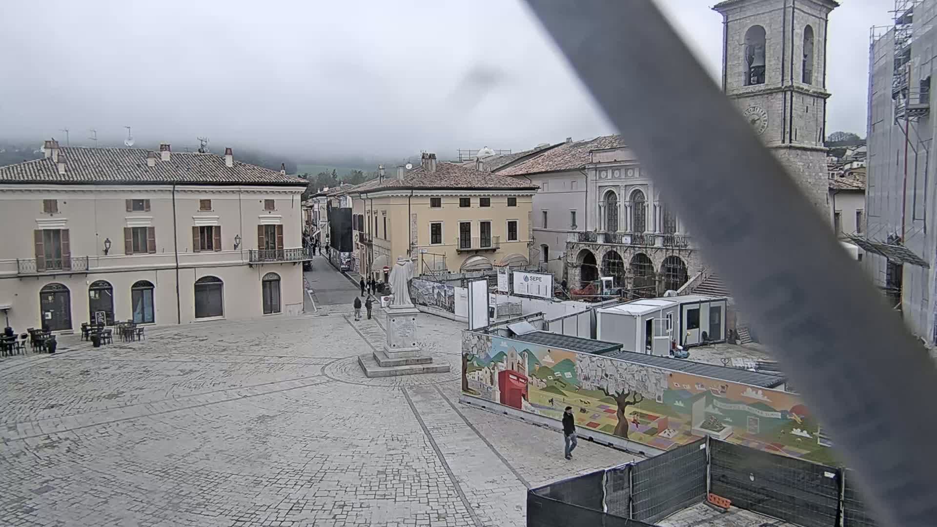 A stone plaza in a town with several buildings, a statue, and construction underway, under overcast skies.