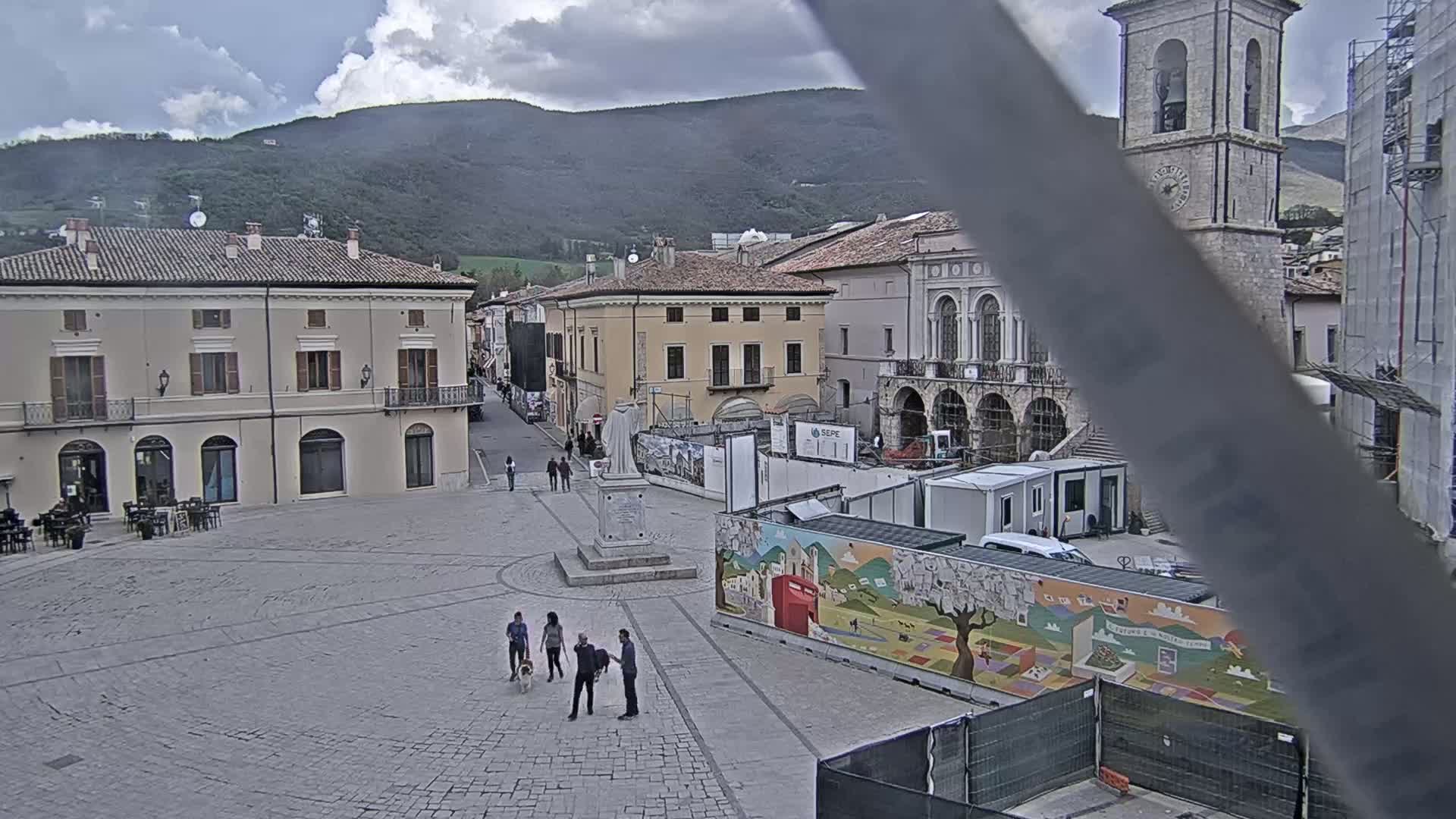 A town square with several buildings, some under construction, is visible under a cloudy sky, with a few people and a dog walking in the square.