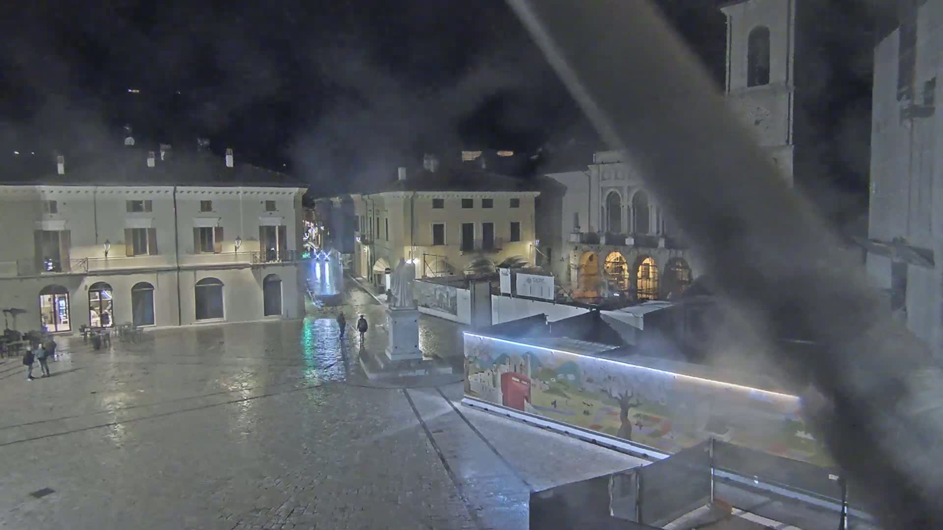 A wet, nighttime town square with several buildings, a statue, and some temporary structures.