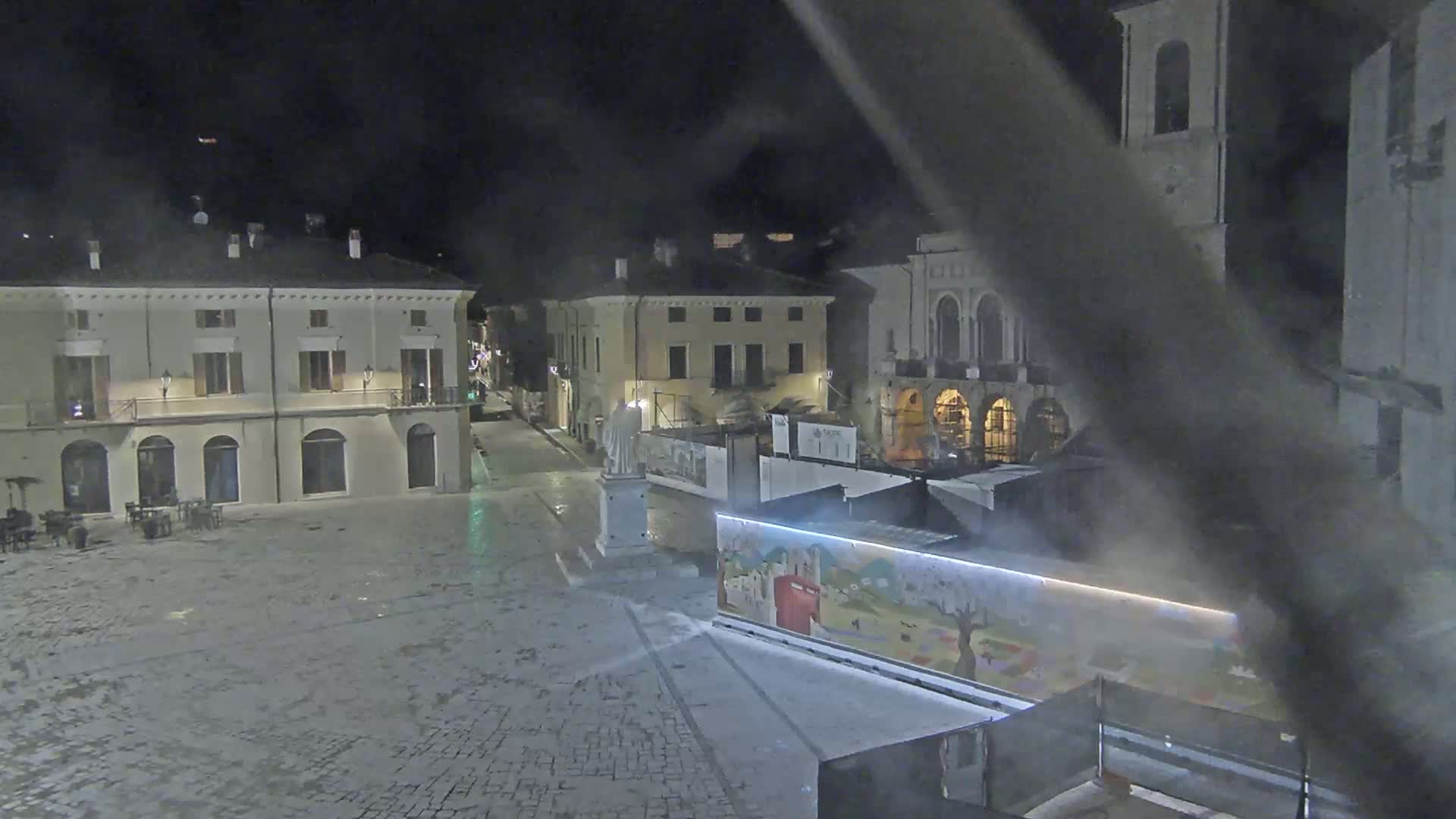 A nighttime view of a town square with several buildings, a statue, and a painted wall partially obscuring construction.