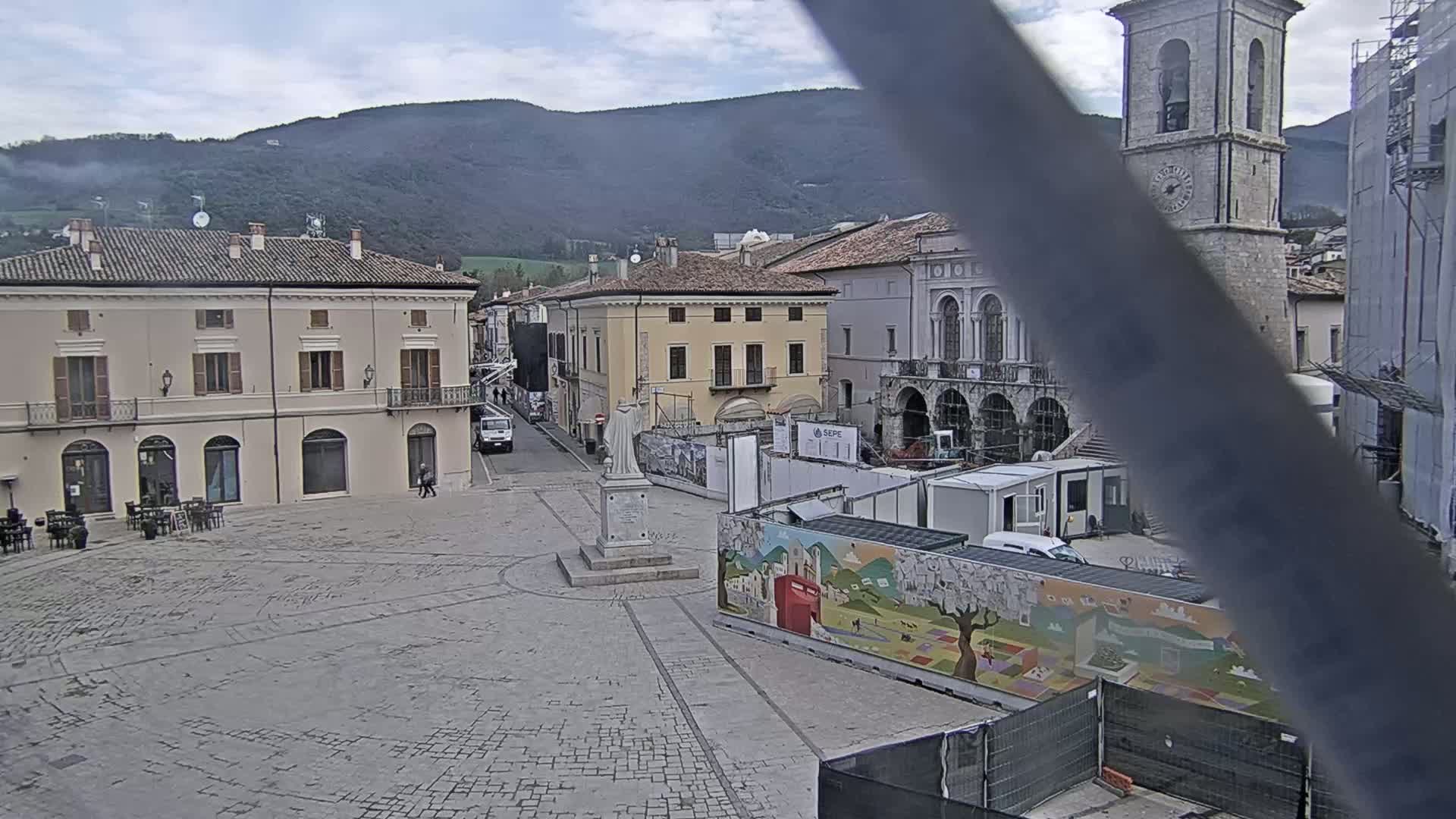 A town square with several buildings, a statue, and construction barriers is visible under an overcast sky with mountains in the background.