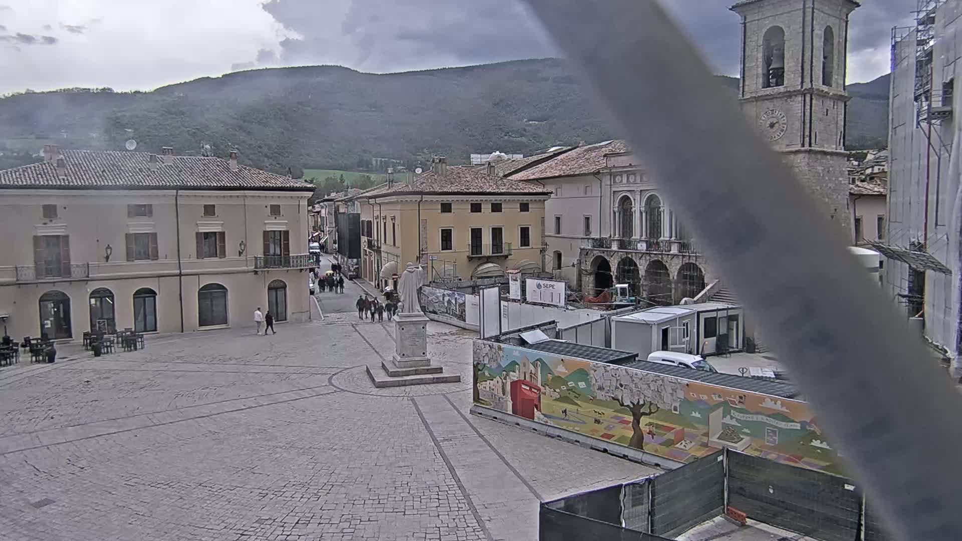 A town square with stone buildings under a cloudy sky, partially obscured by construction and a large mural.