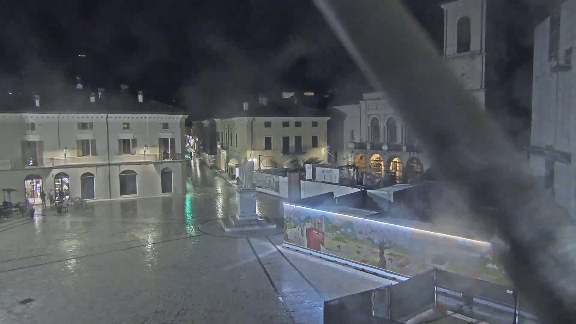 A rain-slicked town square at night, surrounded by historic buildings, with a statue in the center and construction barriers partially obscuring a mural.