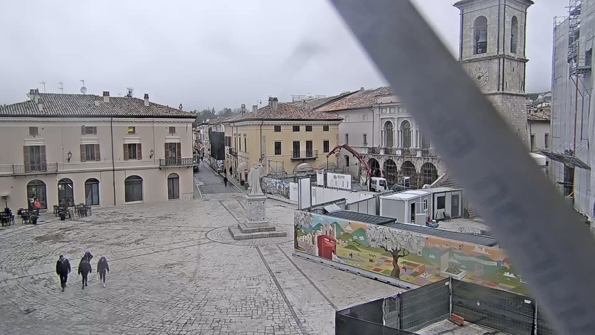 A town square with several buildings, a statue, and construction underway, on an overcast day.