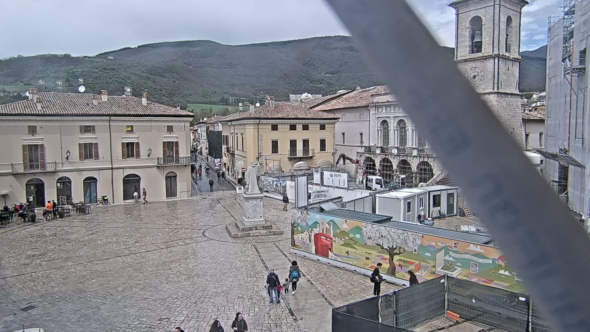 A town square with several buildings, a statue, and construction barriers under an overcast sky.