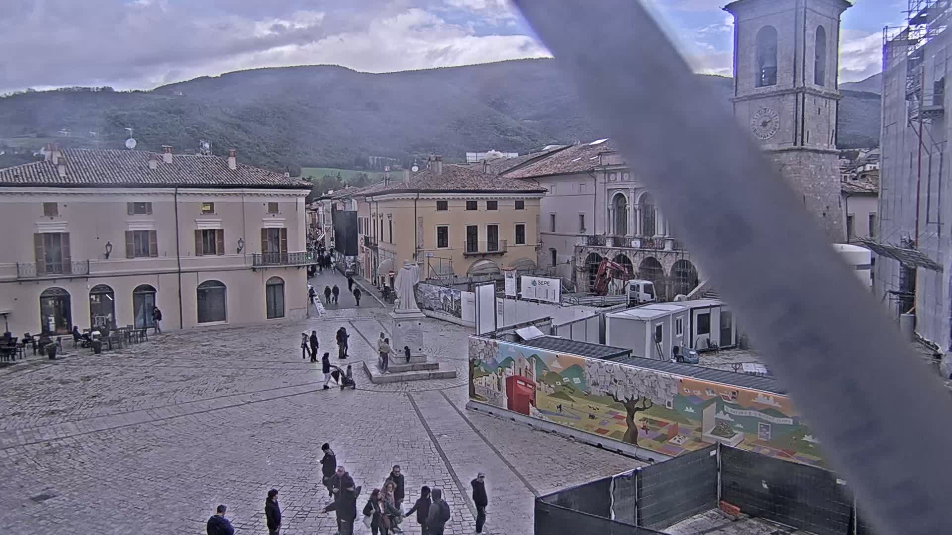 A town square in a mountainous region, under an overcast sky, shows construction in progress near a stone bell tower and several buildings.