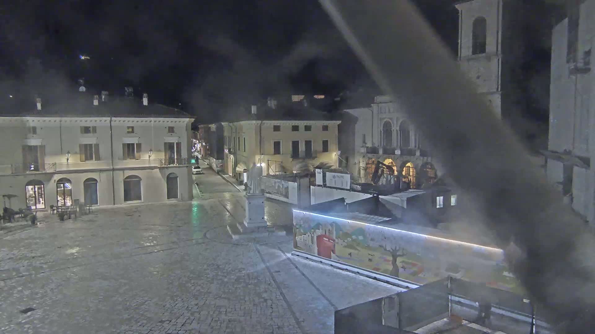 A mostly empty cobblestone town square at night is surrounded by buildings, one of which is under construction, with a statue in the center and a brightly lit mural on a temporary structure.