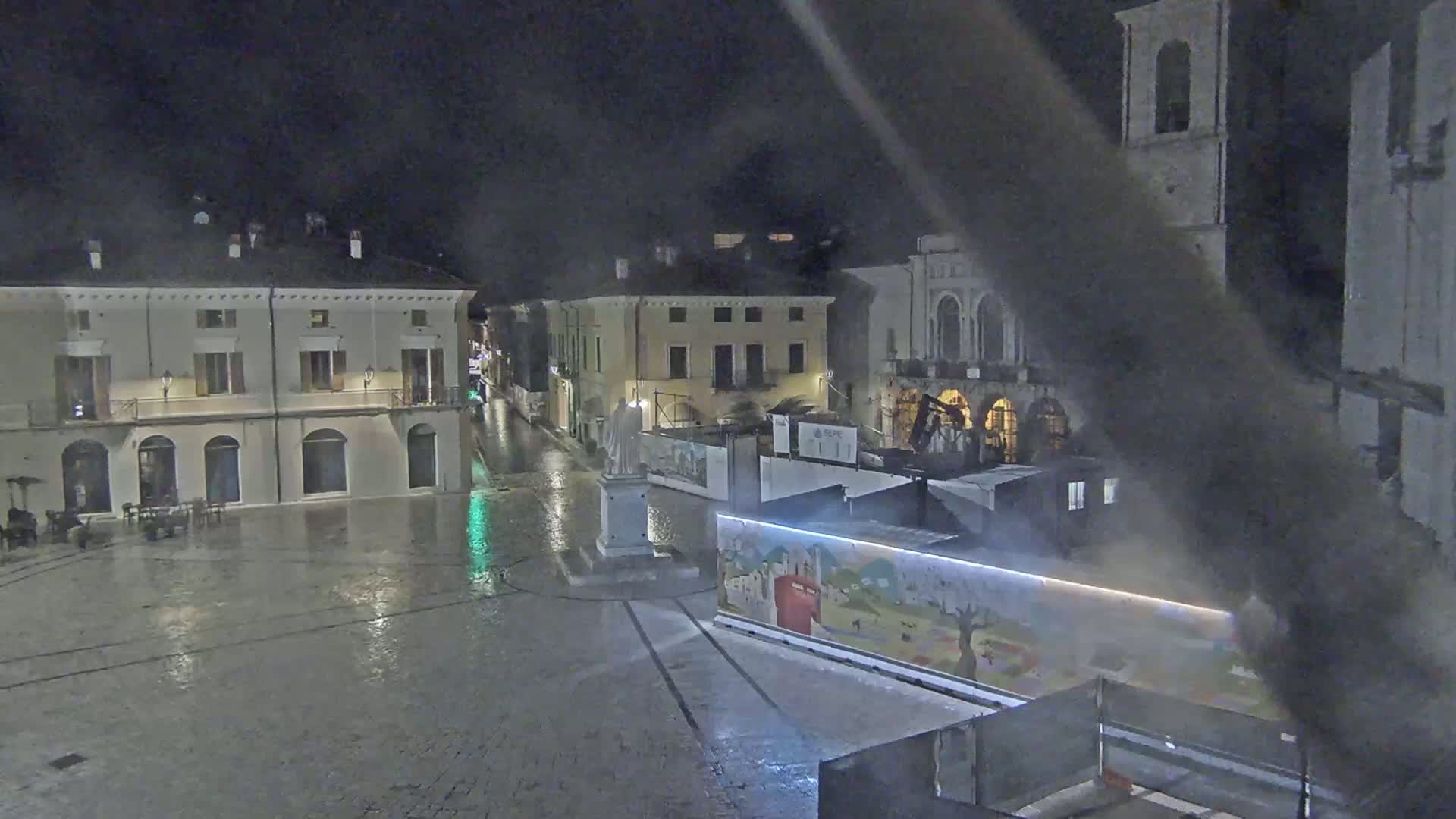 A rain-slicked town square at night, surrounded by buildings, with a statue in the center and construction barriers in the foreground.