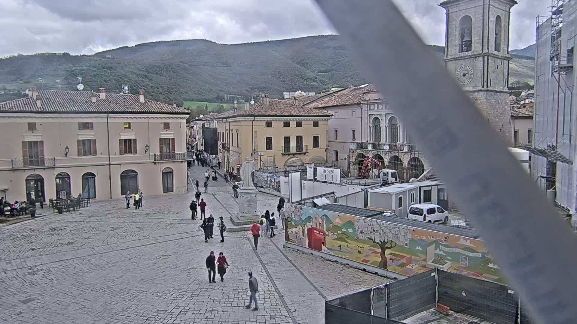 A stone plaza with several people walking around, some buildings, construction, and a backdrop of overcast mountains.