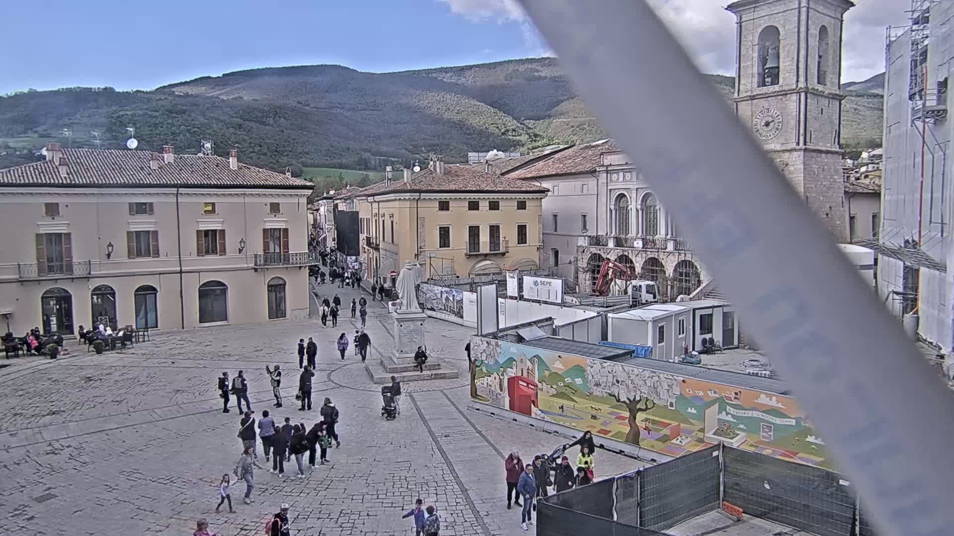 A town square with people walking around, buildings surrounding it, and mountains in the background under a mostly clear sky.