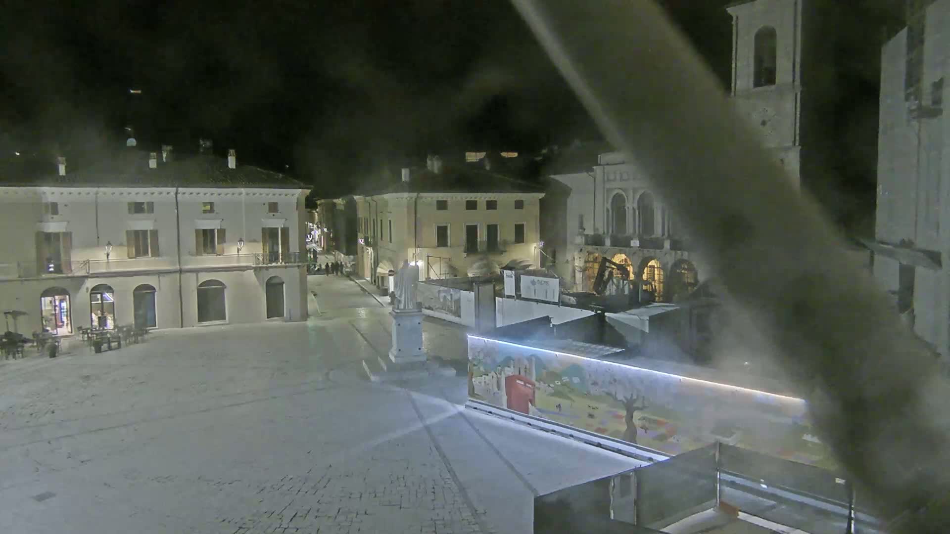 A snow-covered town square at night, surrounded by buildings, with a statue and construction barriers in the center.