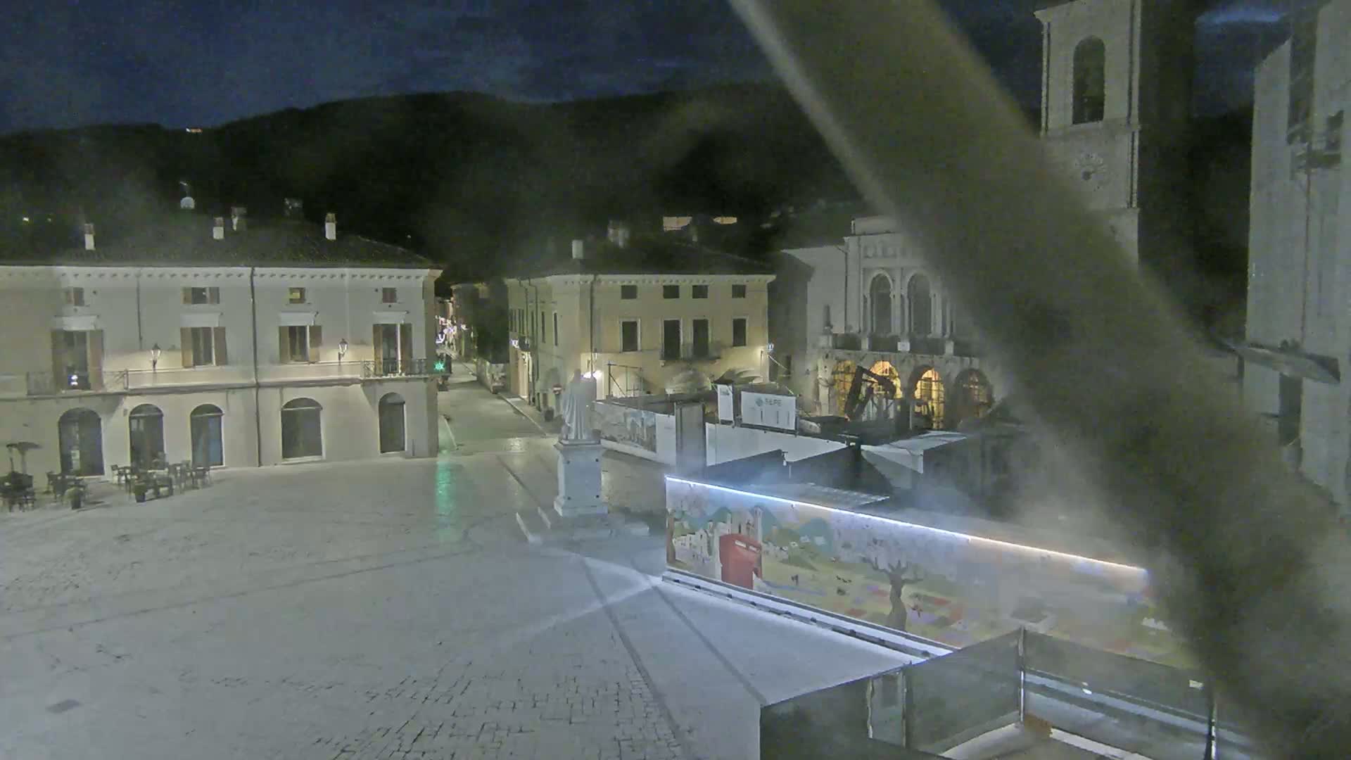 A nighttime view of a town square, featuring several buildings, a statue, and construction barriers, under a dark, possibly overcast sky.