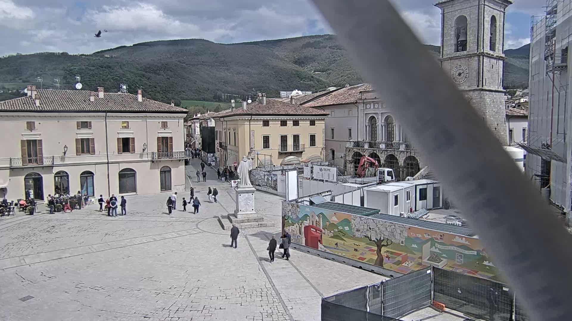 A town square with several buildings, some under construction, is seen under a partly cloudy sky, with a bird flying overhead and mountains in the background.