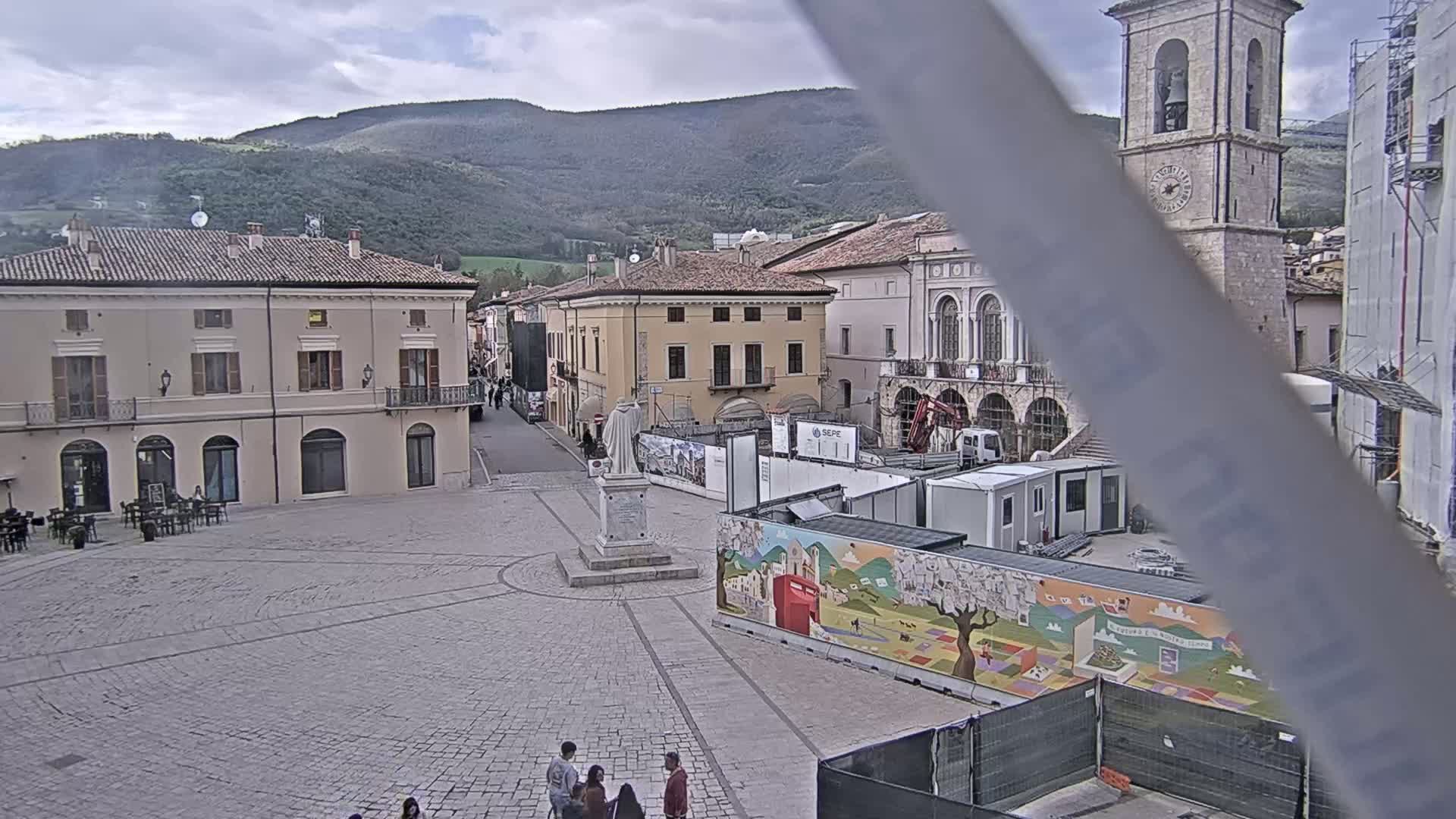 A town square, partially under construction, is situated in front of several buildings under a cloudy sky, with mountains visible in the background.